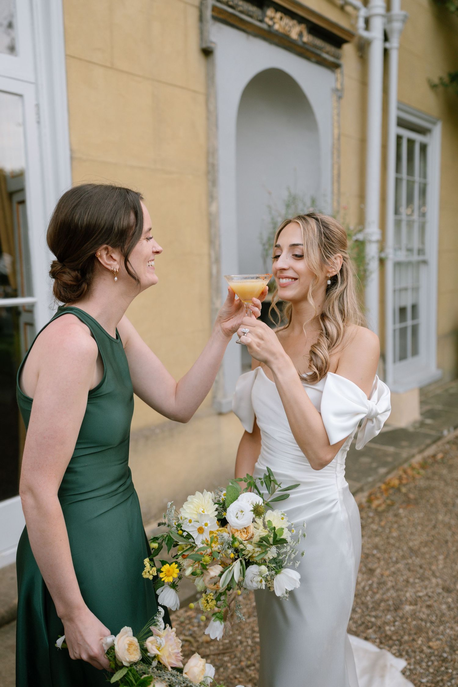 Two women in green and white dresses share a joyful moment while holding bouquets at an outdoor event.