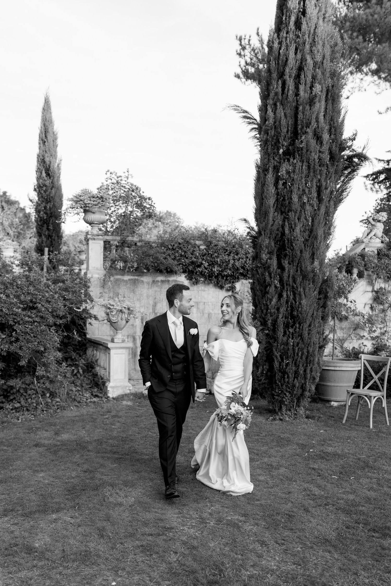 Black and white photo of couple walking on lawn with cypress trees in background.