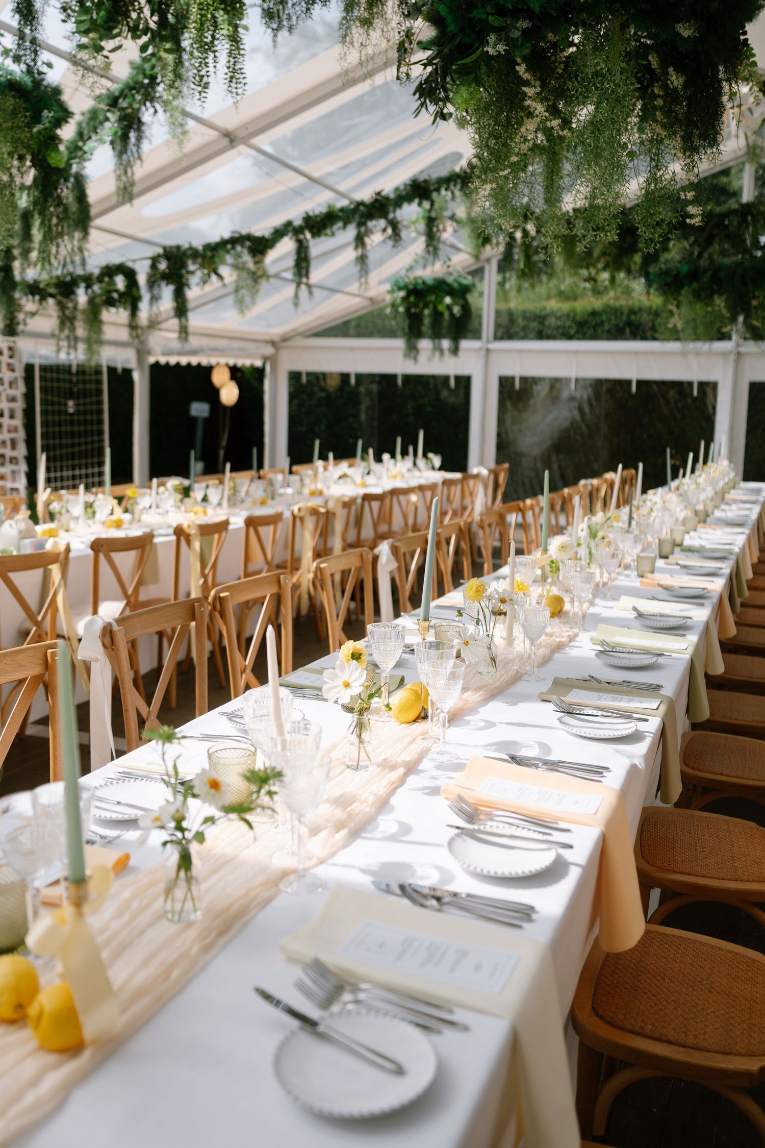 Elegant wedding reception tables set with white linens and wooden cross-back chairs under a clear tent.