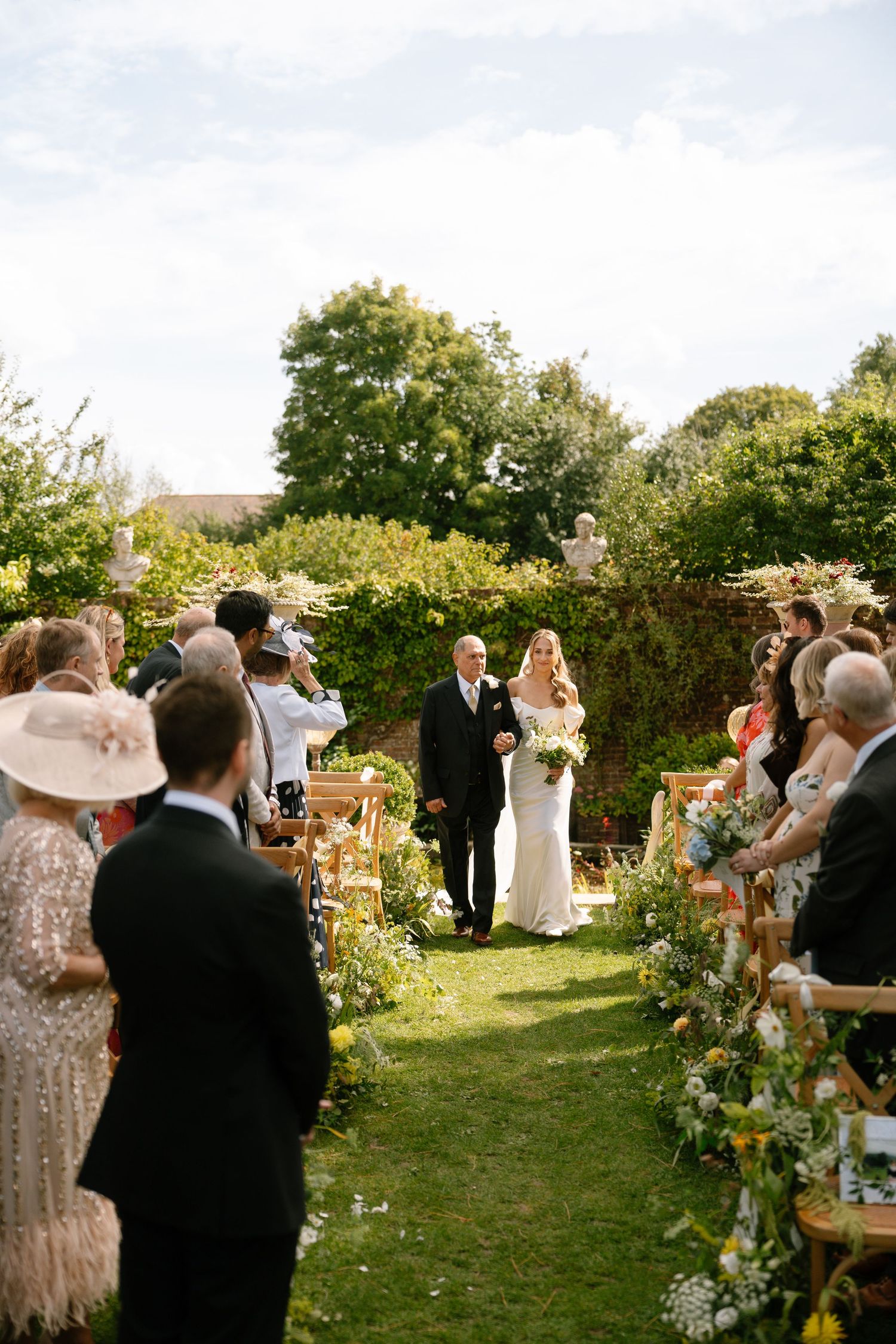 Garden wedding ceremony with guests gathered around floral decorations and wooden chairs.