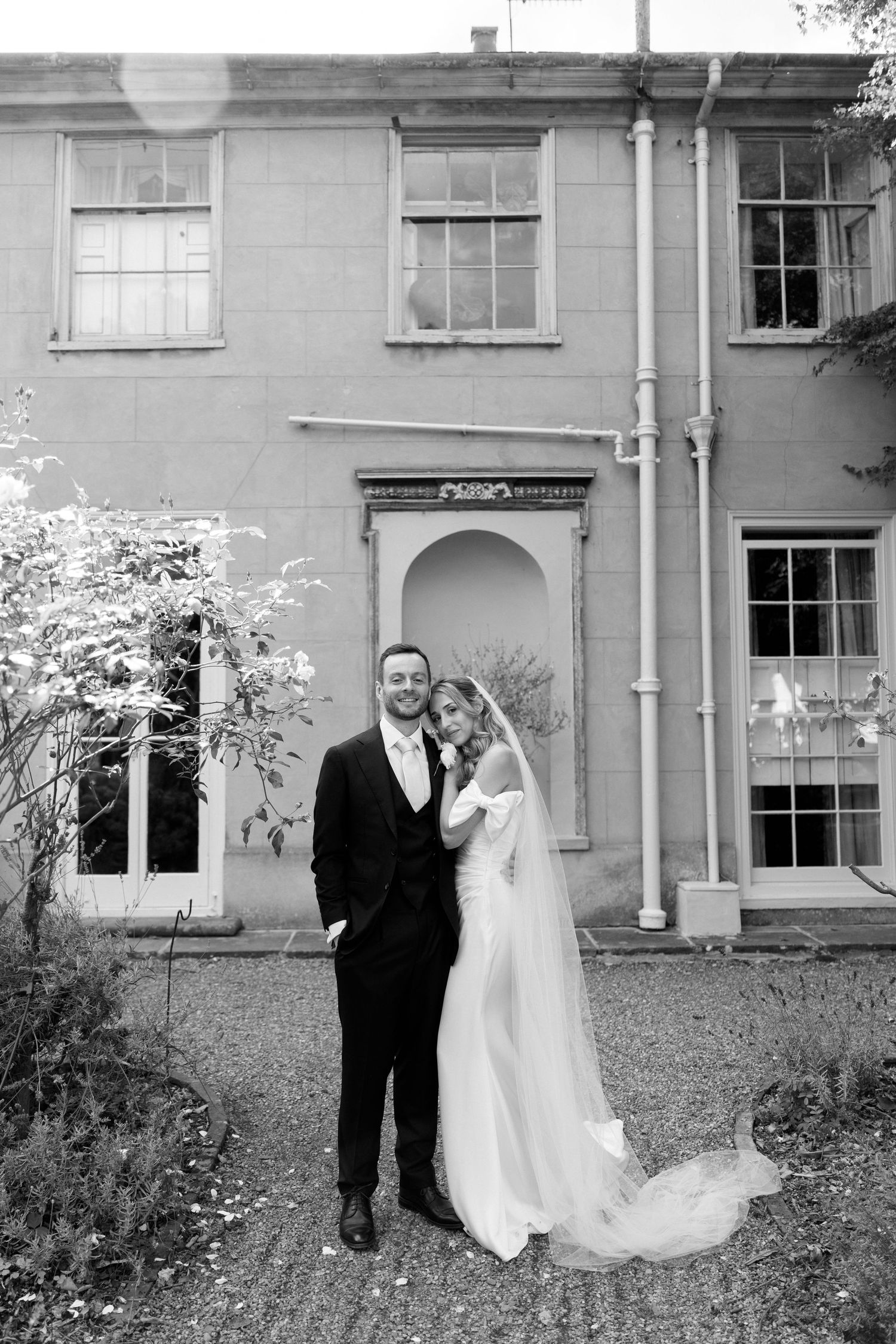 Black and white wedding portrait against classic architecture with garden elements.