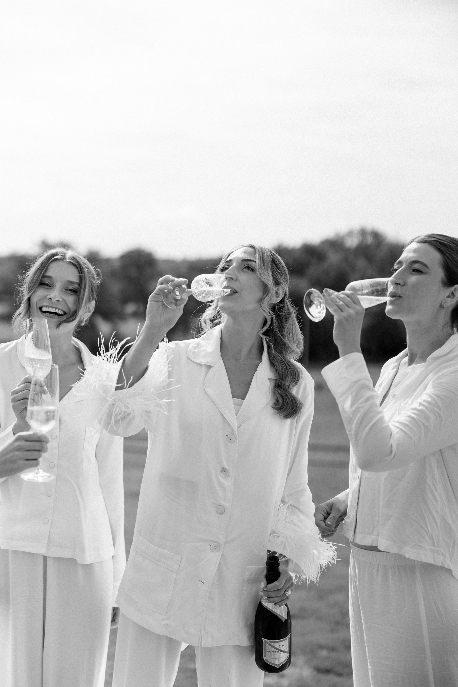 Three people in white outfits share champagne and laughter during an outdoor celebration.