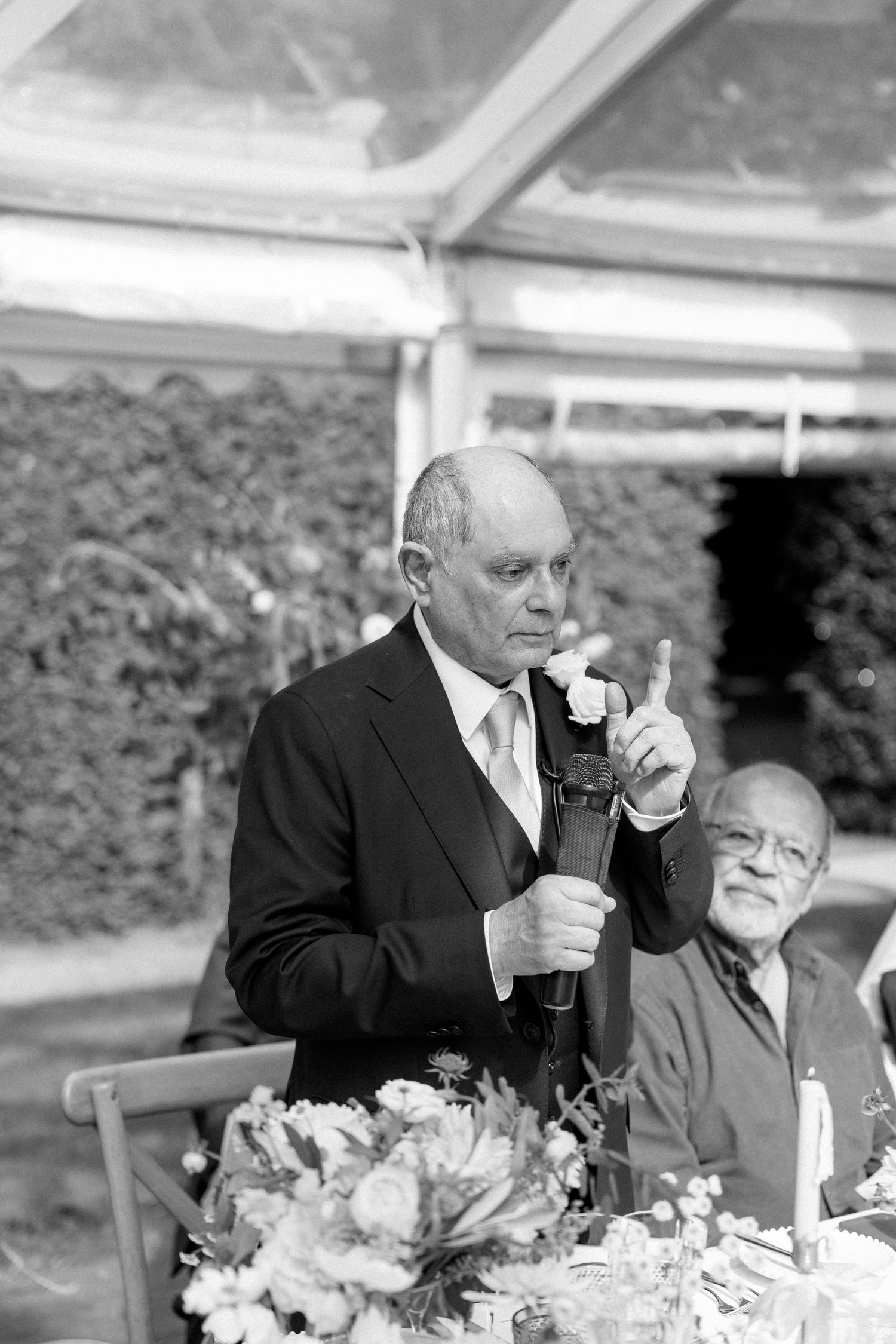 A formal speaker delivers remarks at an elegant wedding reception with floral centerpieces.