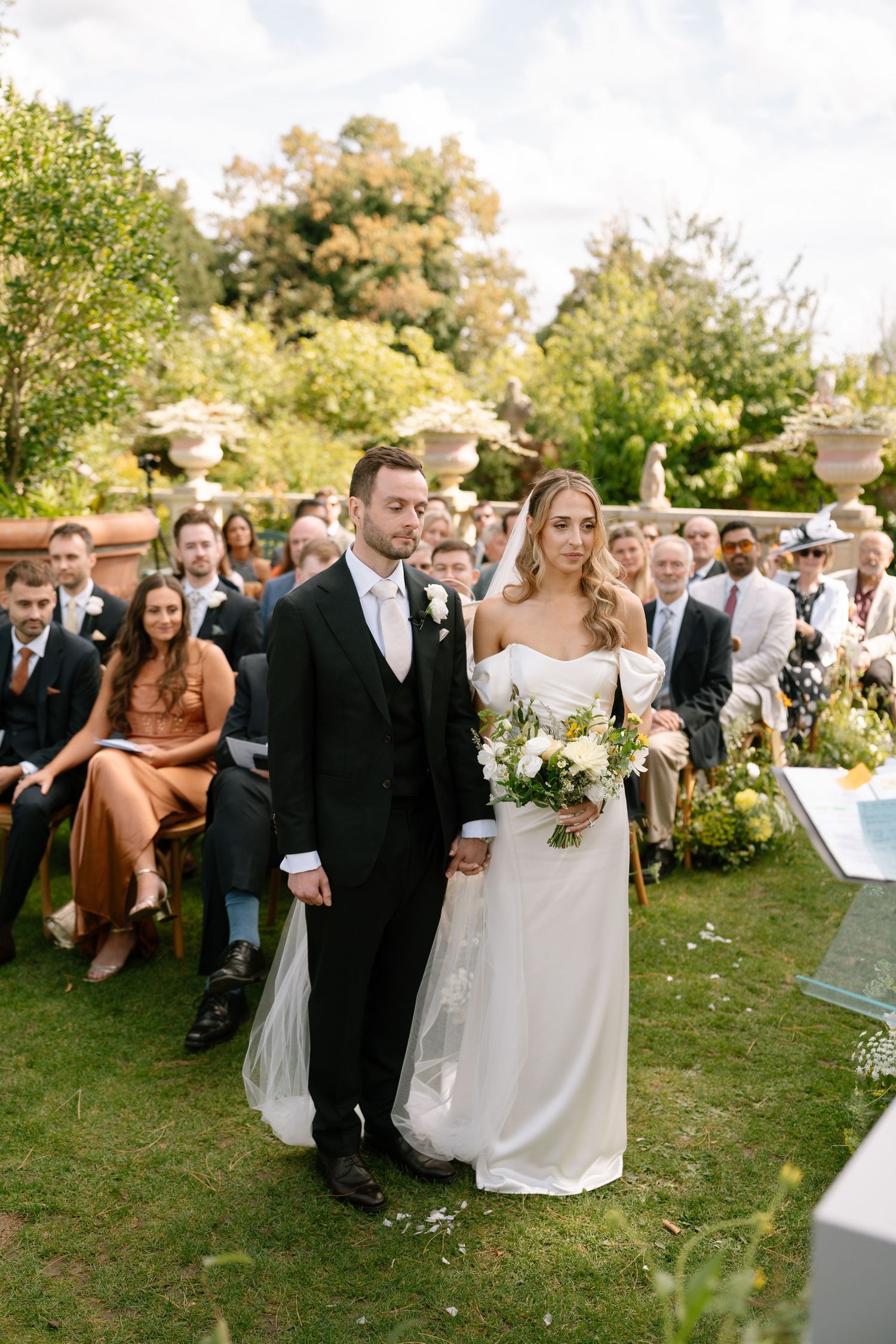 Wedding processional moves through an elegant garden setting with guests watching from their seats.