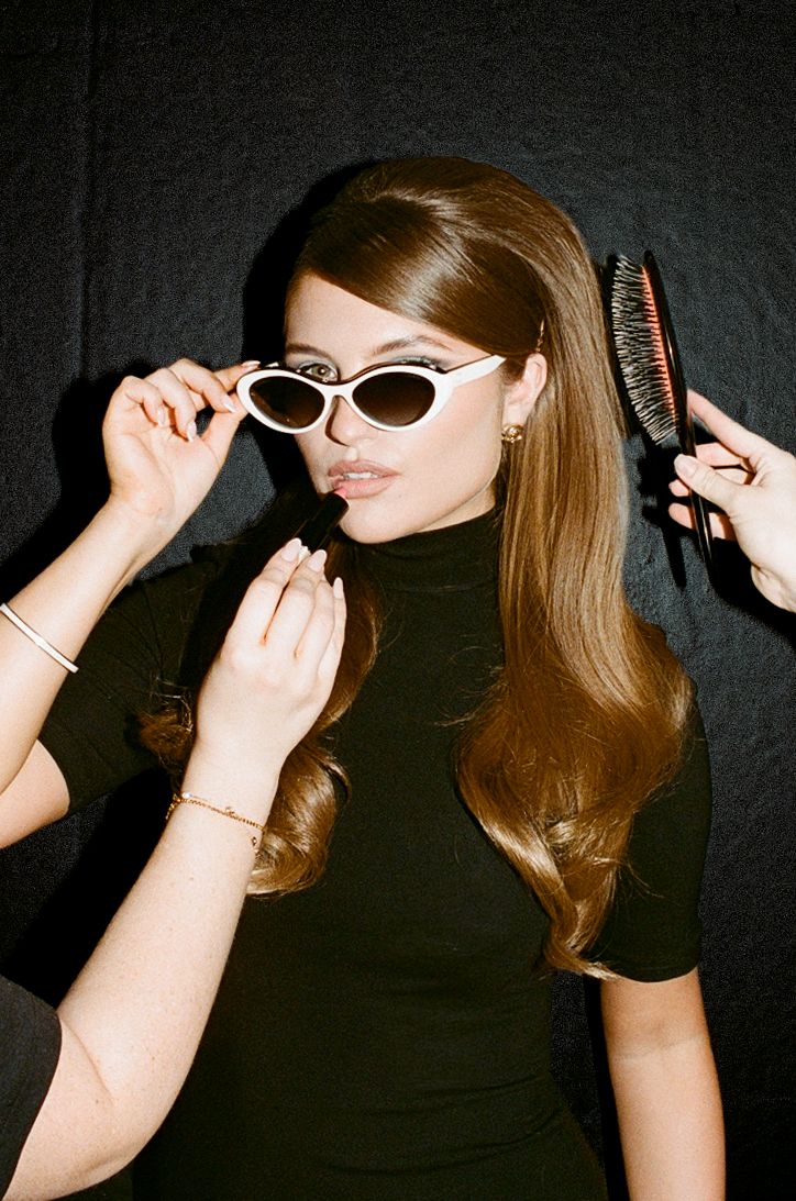 A woman in a black turtleneck adjusts white cat-eye sunglasses while getting hair and makeup done backstage.