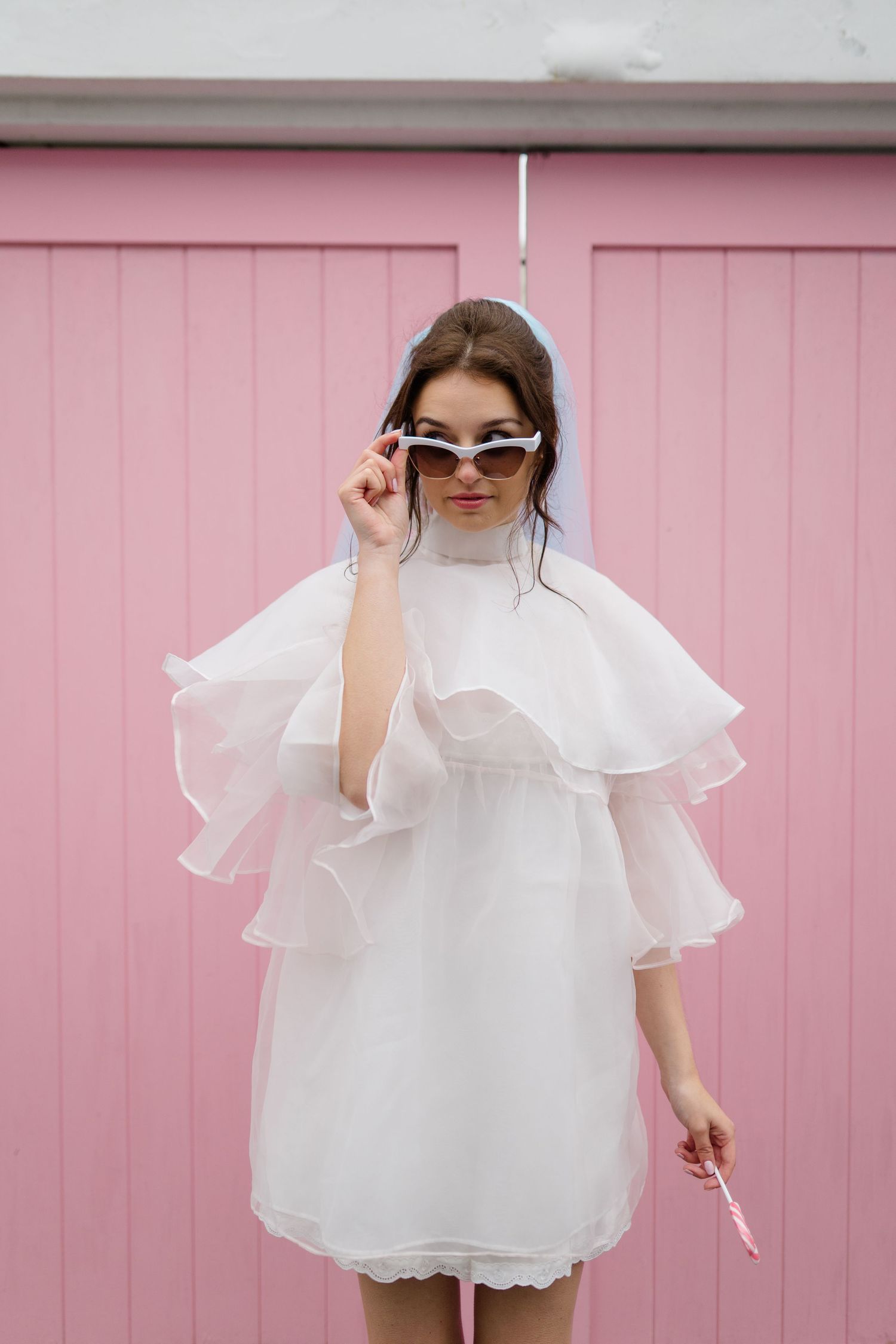 Woman in white ruffled mini dress and white cat-eye sunglasses posing against a pink wall.