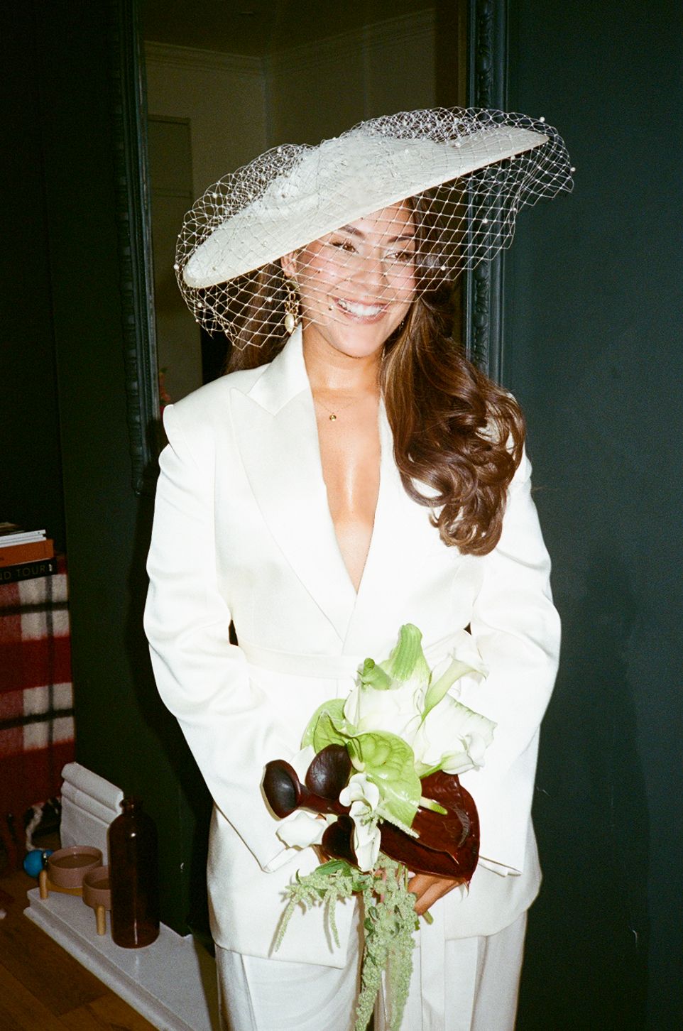 Smiling bride in white suit and wide-brim hat with veil, holding a green floral bouquet indoors.
