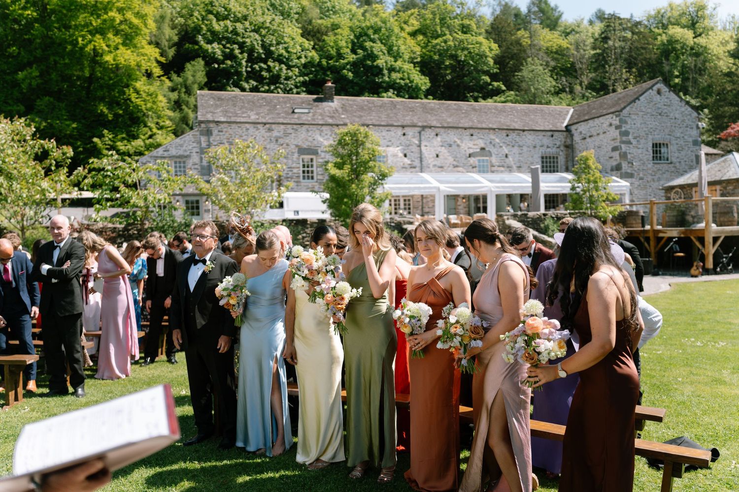 Wedding guests gather outdoors on a sunny day with stone buildings in the background.
