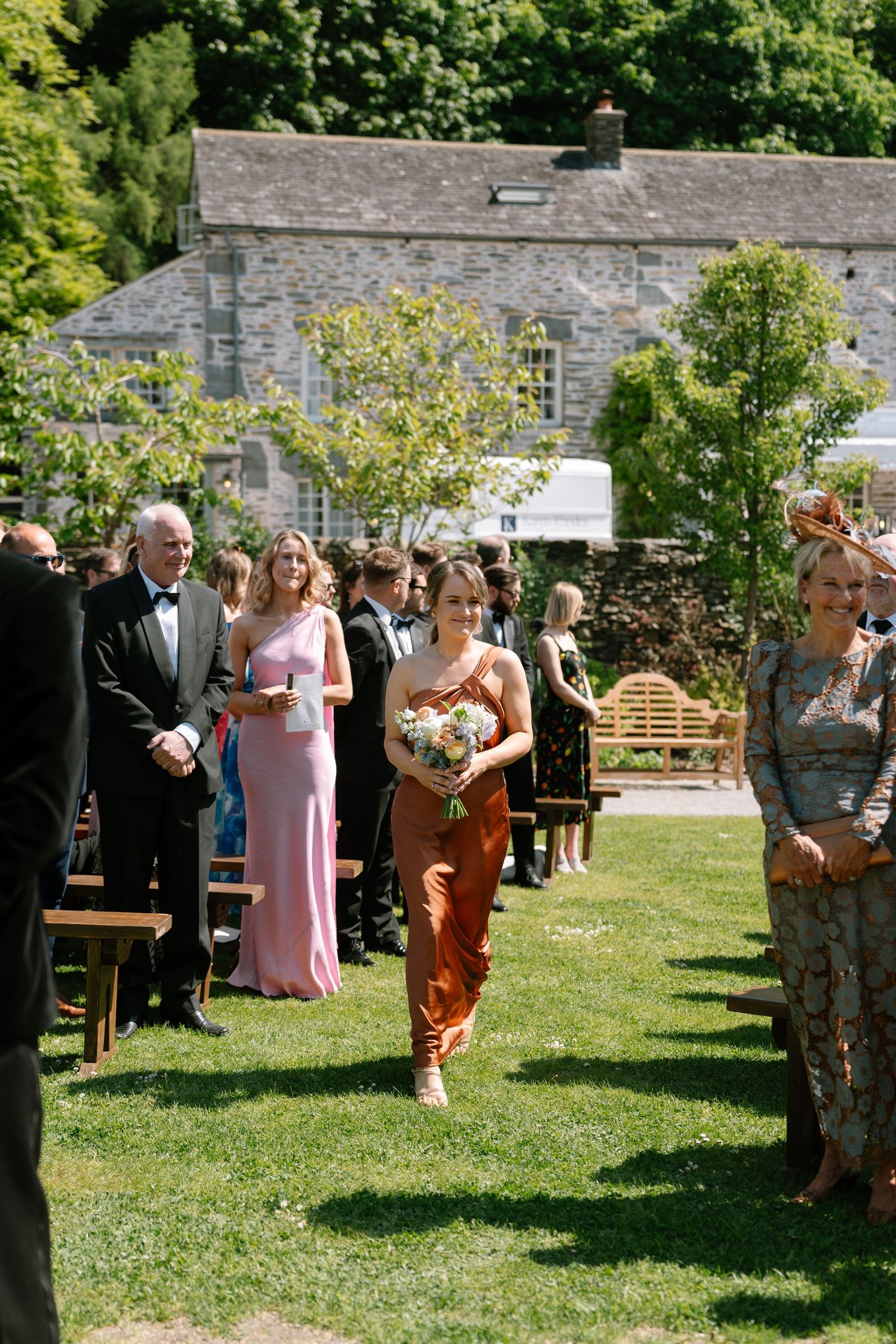 Wedding party and guests mingle on a sunny lawn with stone buildings in the background.