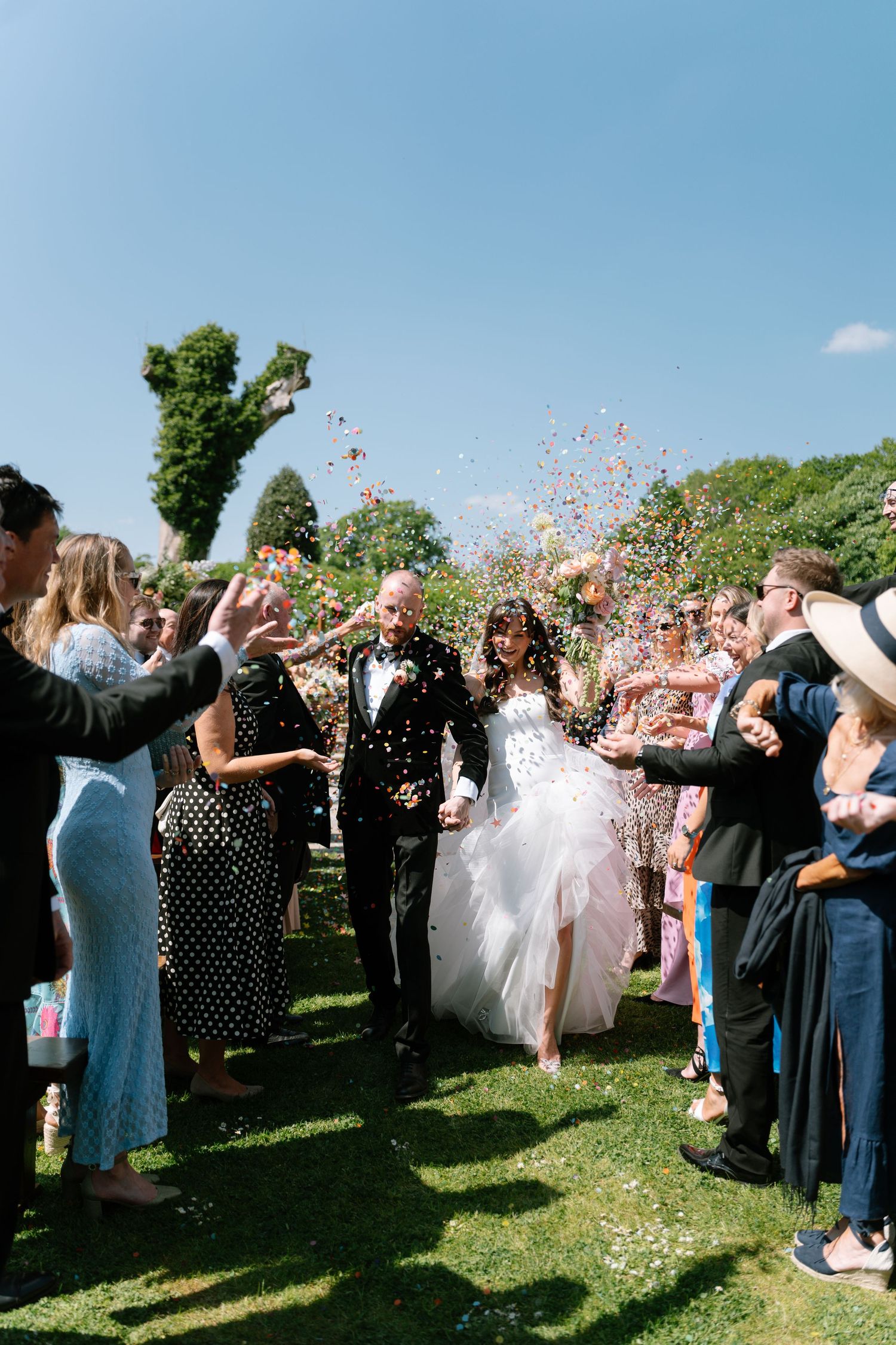 Wedding guests throw confetti at a joyous outdoor celebration on a sunny day.