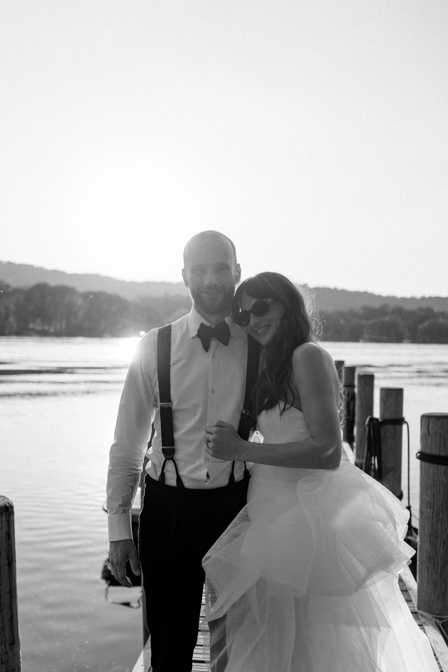 Couple in wedding attire walking along a wooden dock at sunset by the lake.