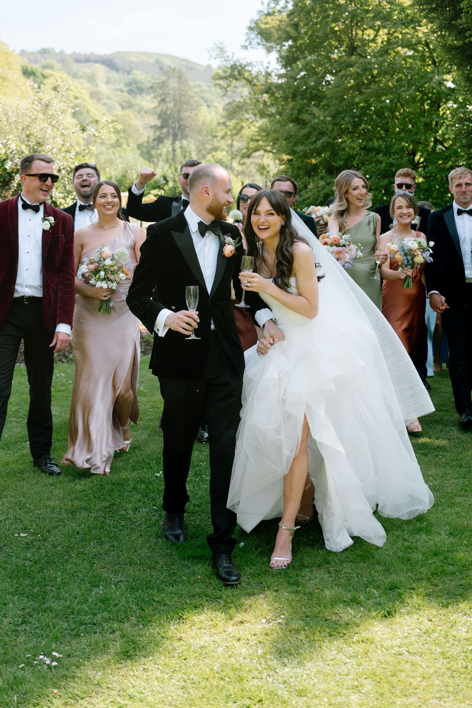 Wedding party celebrates on a sunny lawn with mountains in background as newlyweds share a joyful moment together.