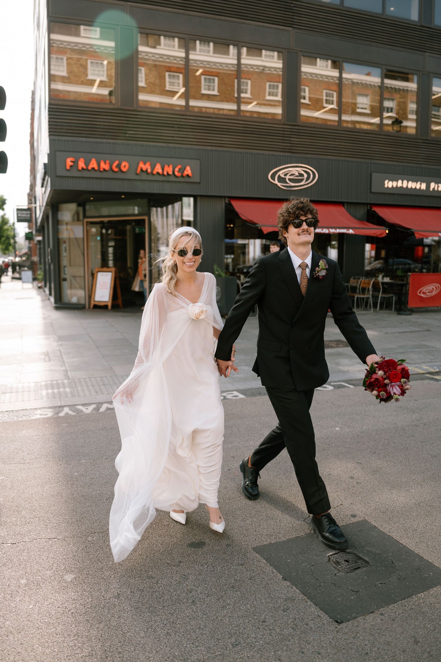 A couple in wedding attire walks past Franco Manca restaurant in an urban setting.