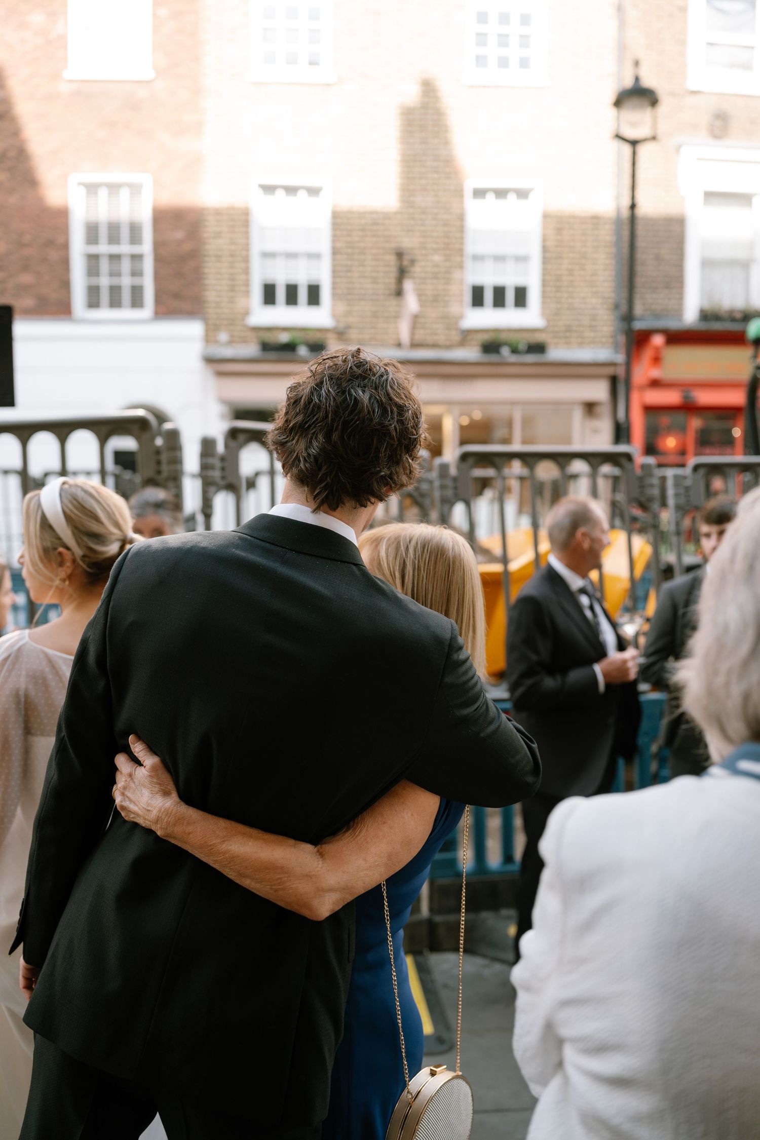 A couple embraces while surrounded by wedding guests on an outdoor terrace.