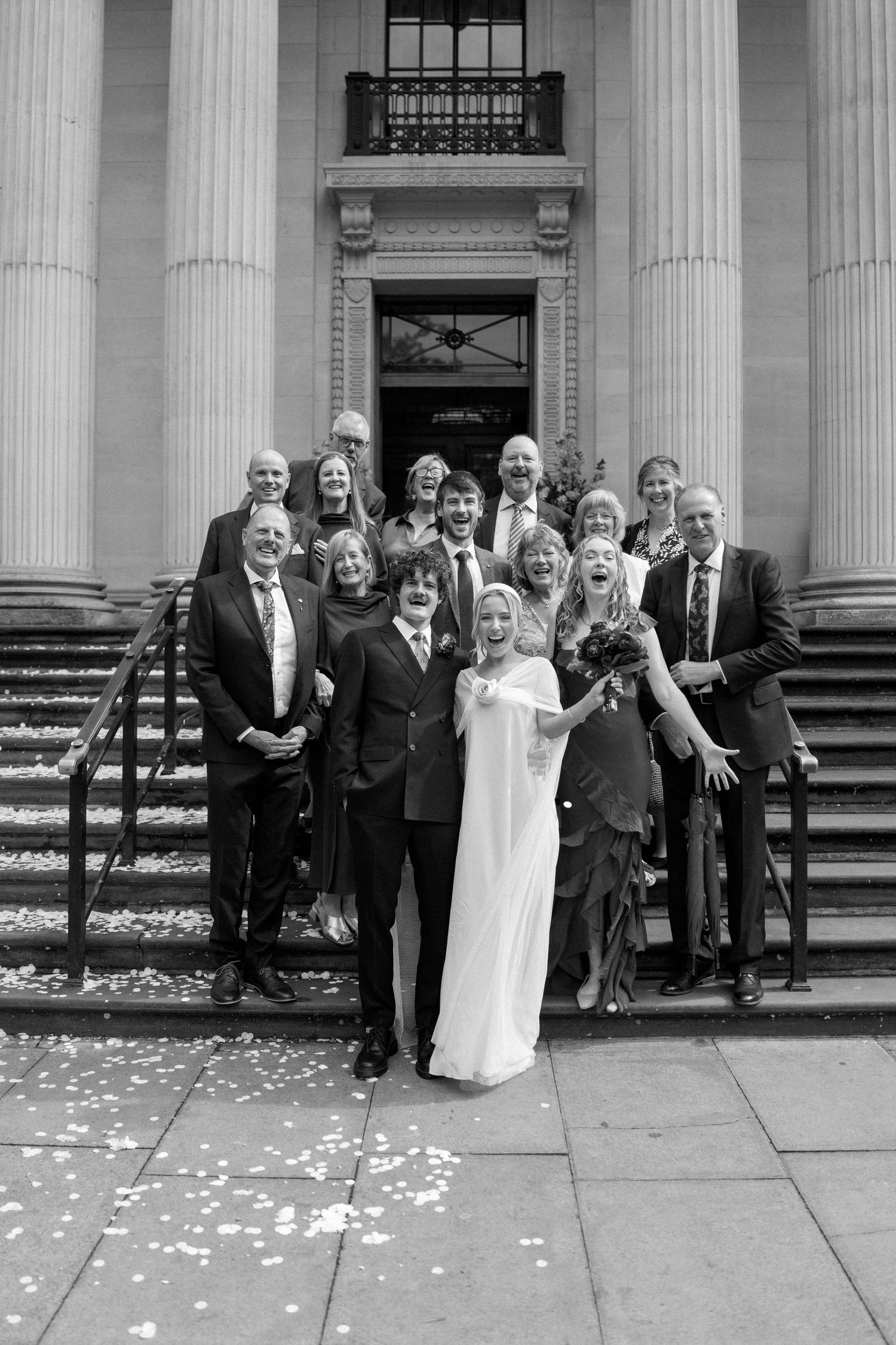 Wedding group poses for photo outside classical building with white columns.