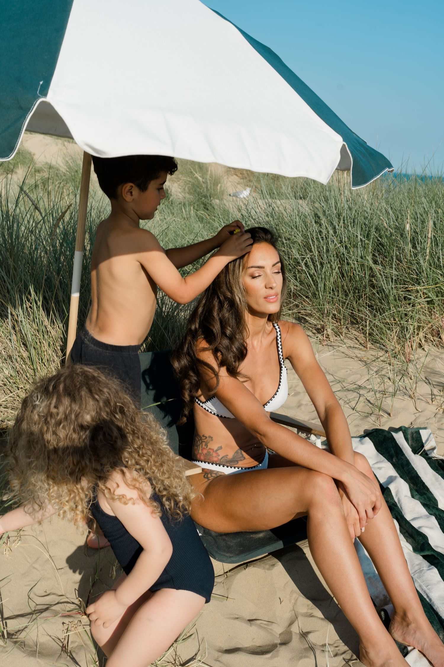 People lounging on striped beach towel under white umbrella on sandy dunes.