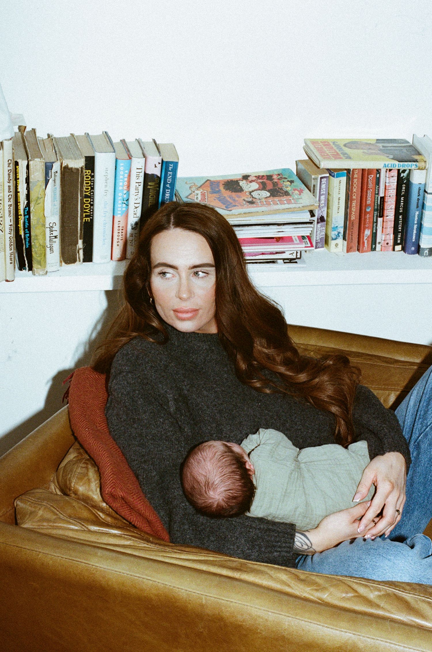 Woman with long auburn hair holding a newborn baby on a leather sofa with bookshelves in background.