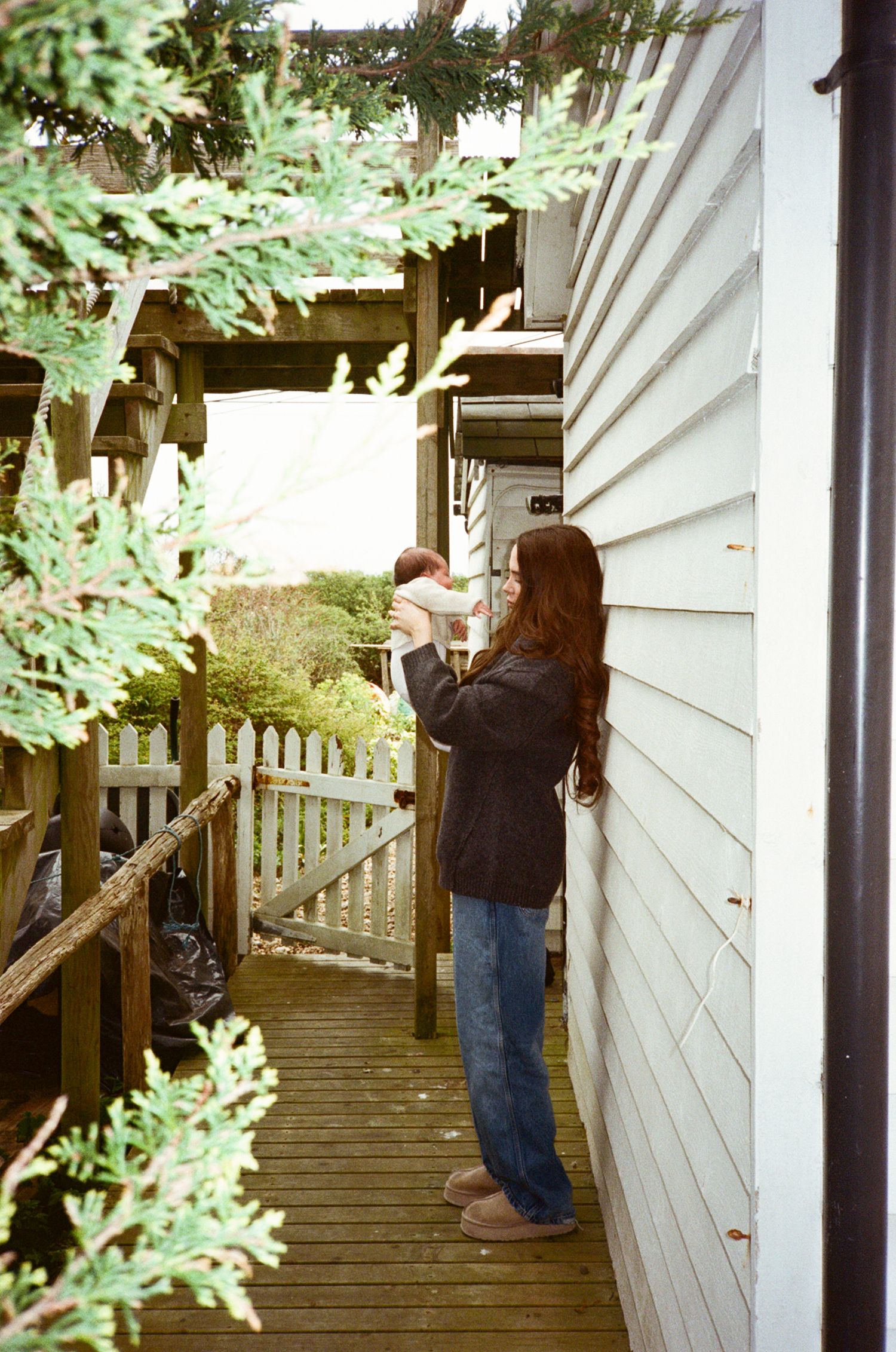 A woman with long hair holds a baby on a wooden deck porch beside a white clapboard house surrounded by greenery.