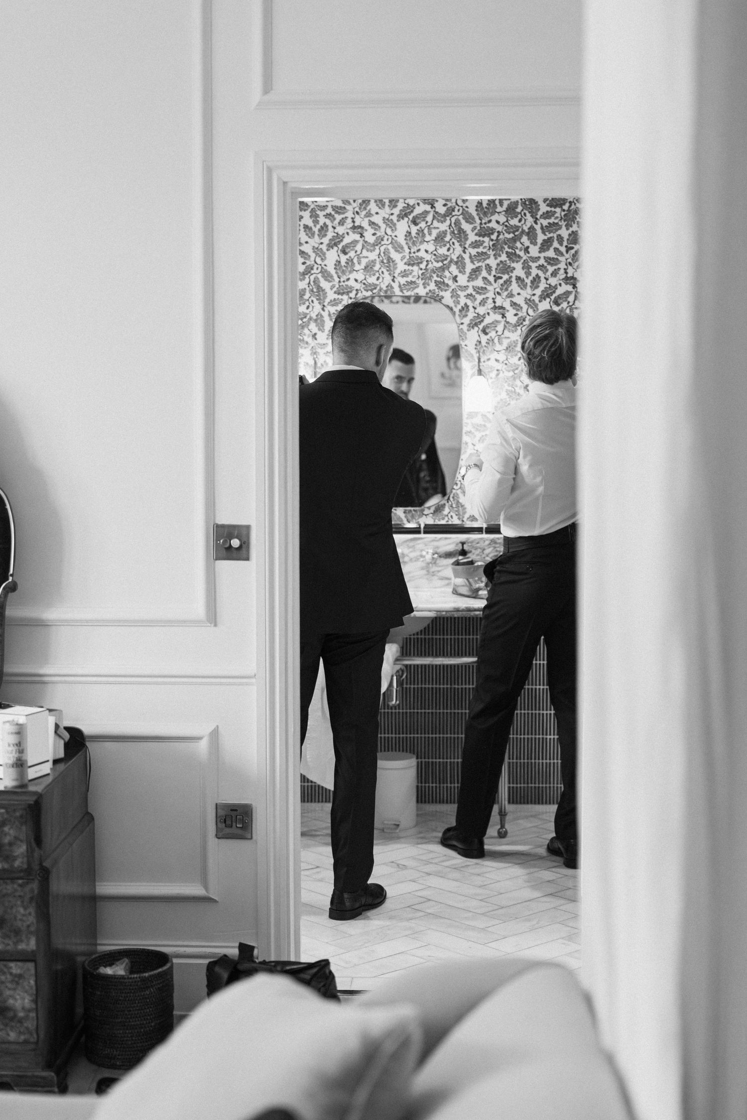 Black and white photo of groomsmen getting ready in elegant bathroom with patterned wallpaper.