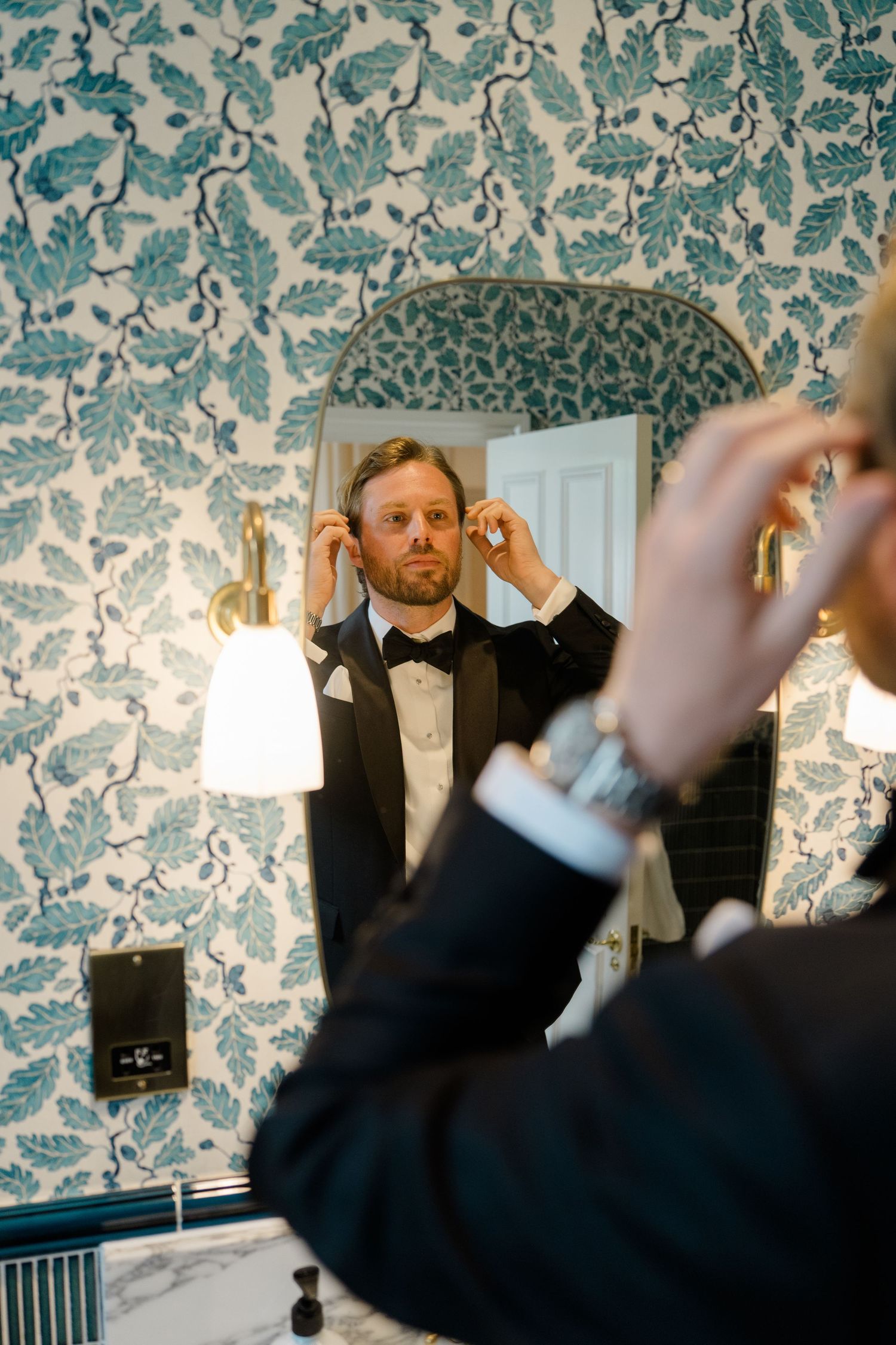 Groom in black tuxedo adjusting his bow tie in a mirror with blue floral wallpaper backdrop.