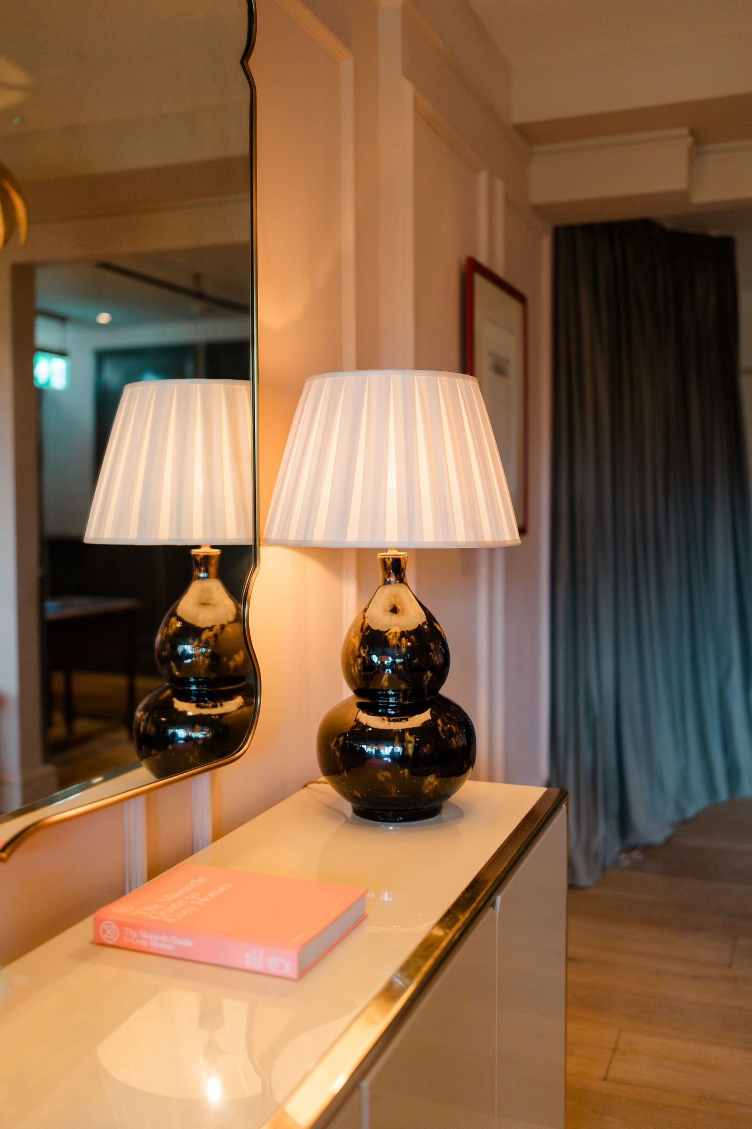 Elegant console table with black gourd-shaped lamps, white pleated shades, and a pink book beside a gold-framed mirror.