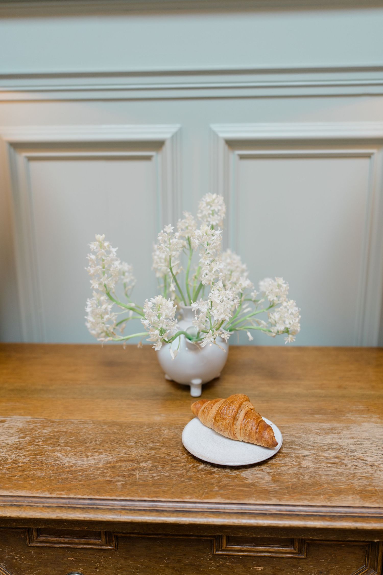 A golden croissant on a white plate beside a white vase of delicate white flowers on a wooden sideboard.