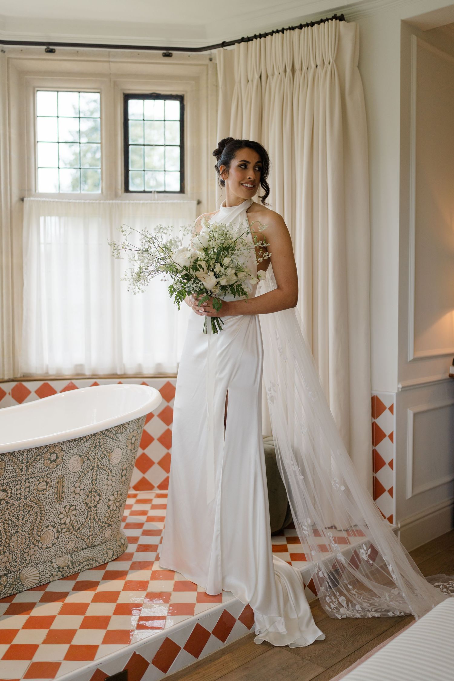 Radiant bride in halter neck gown holding white bouquet standing in ornate tiled bathroom.