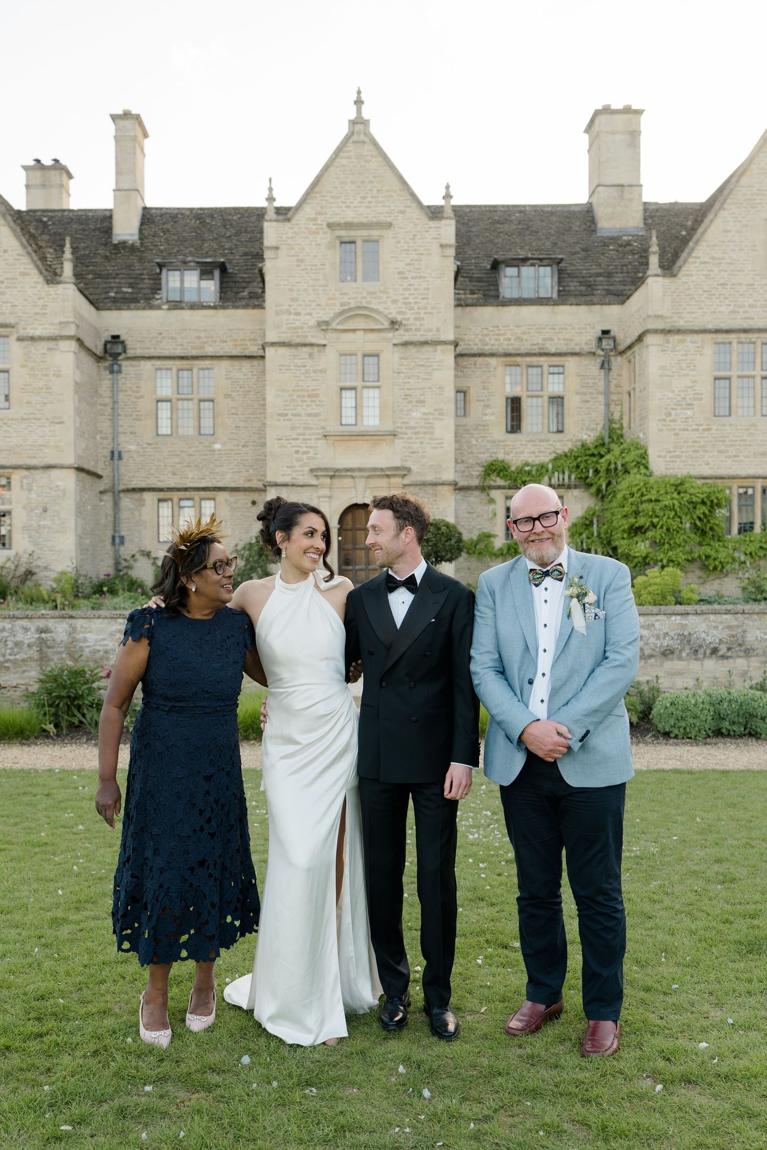 Bride and groom pose with two guests in front of grand Cotswolds stone manor house at dusk.