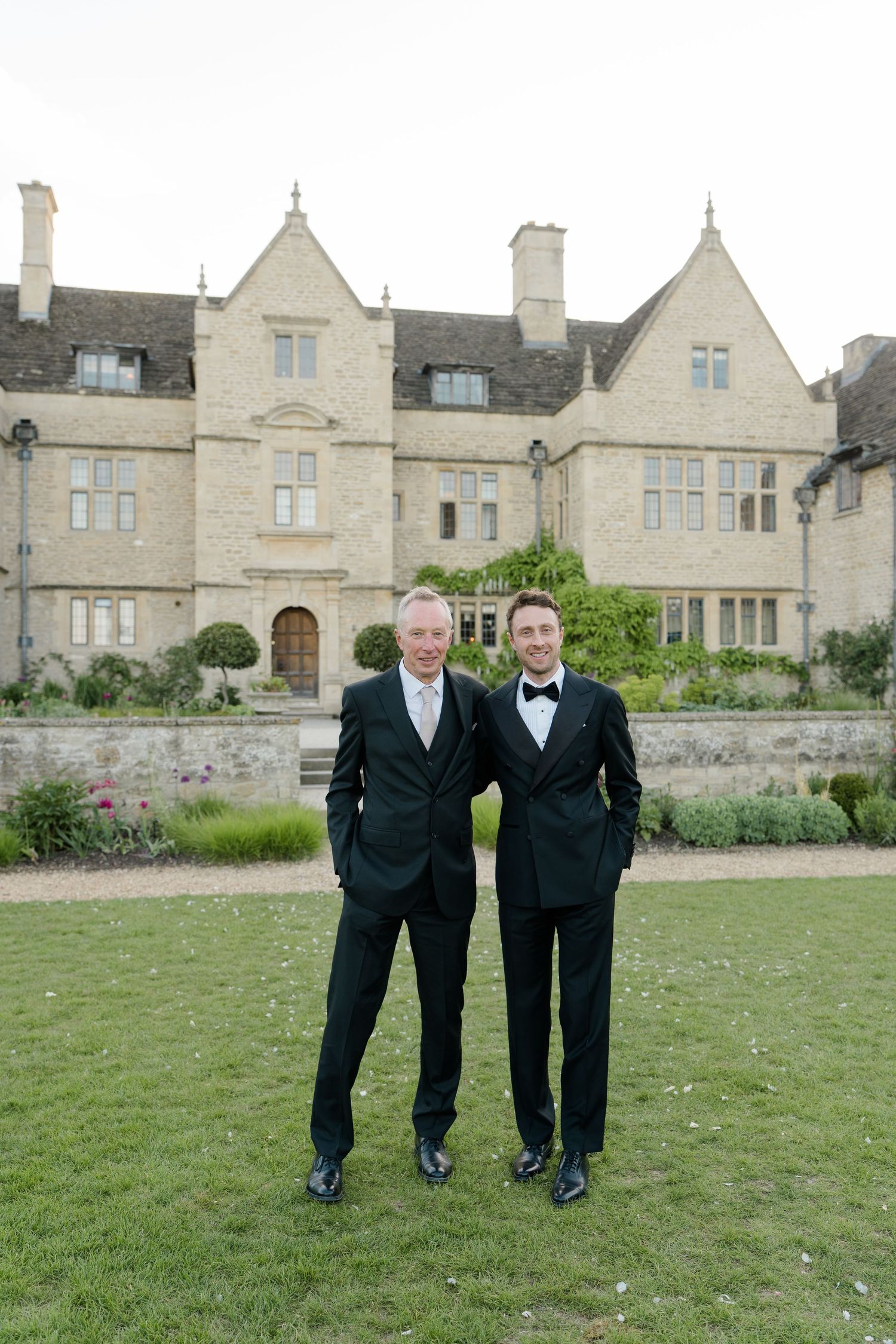 Father and son in matching black tuxedos pose arm in arm before a grand English manor house.