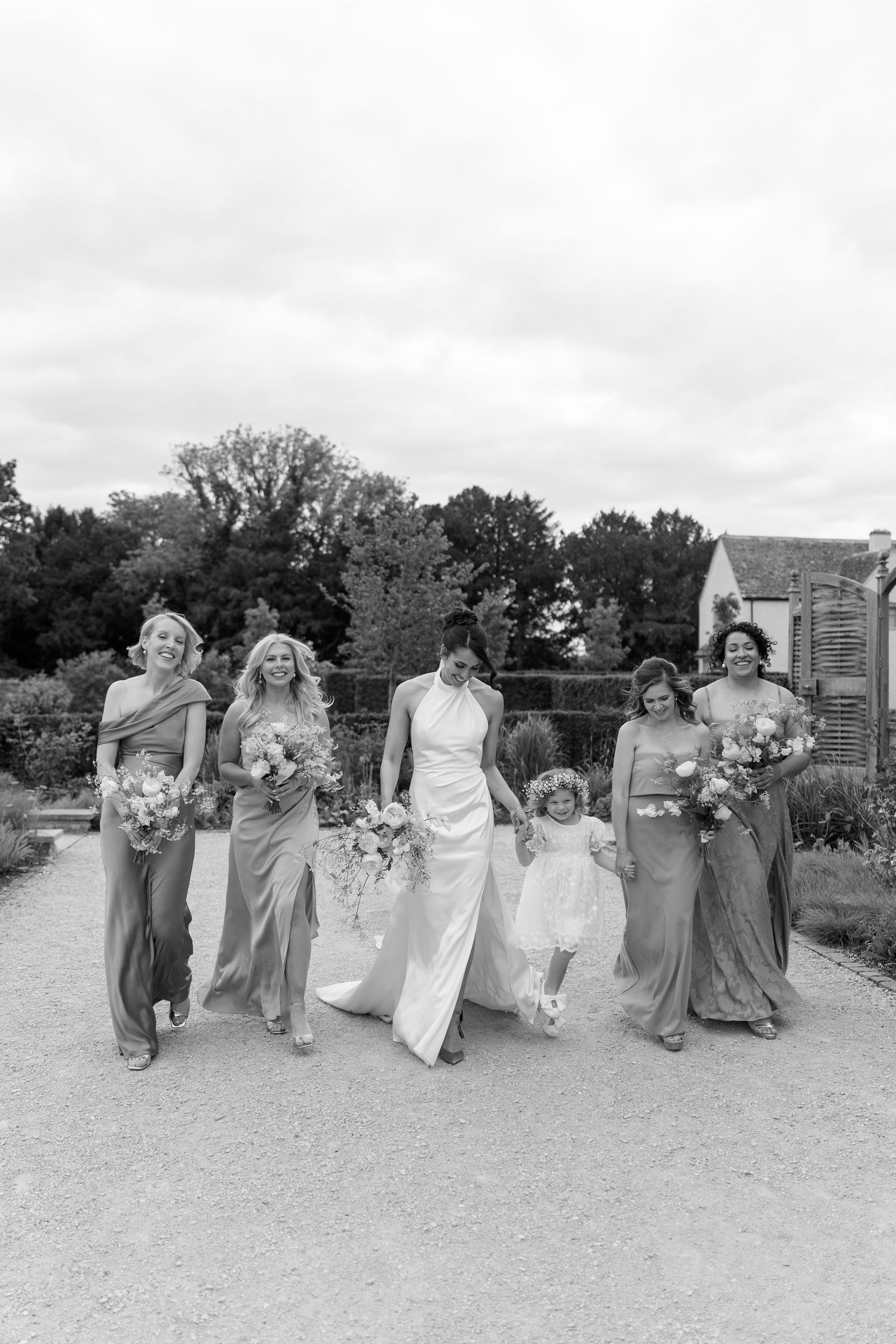 Bride walks with bridesmaids in elegant gowns and a flower girl on a garden path in black and white.