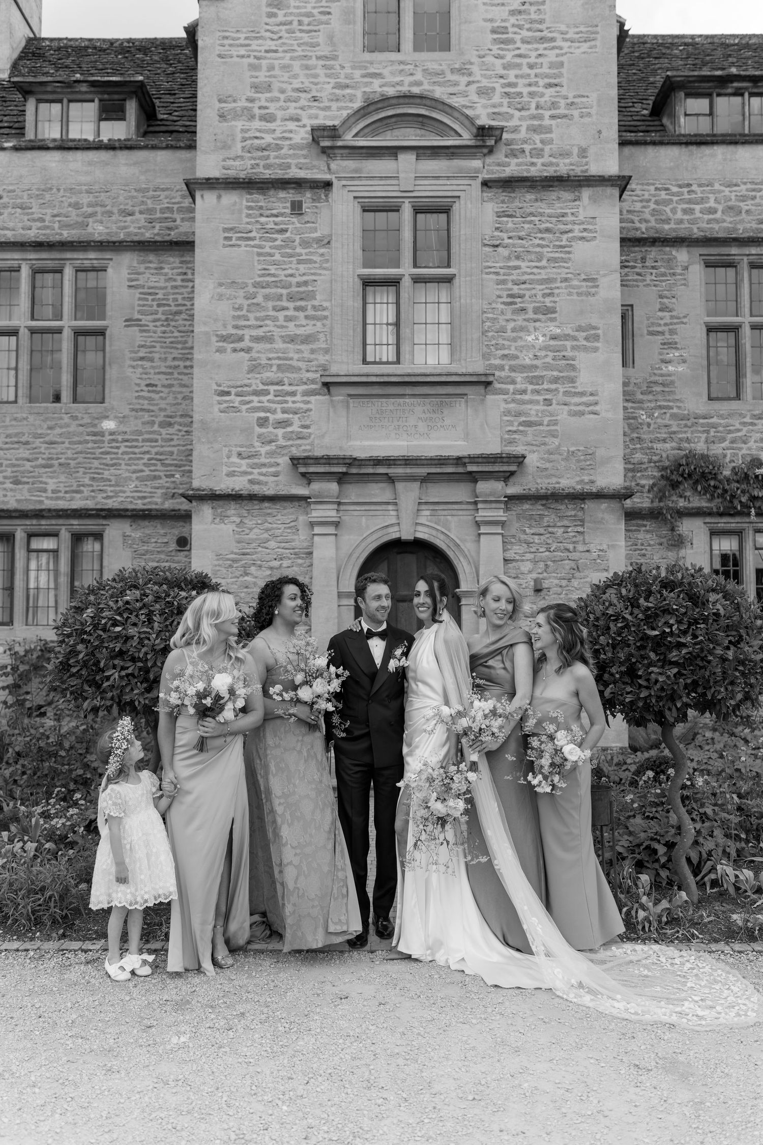 Bride, groom, and bridesmaids in sage dresses pose before historic stone manor in black and white.