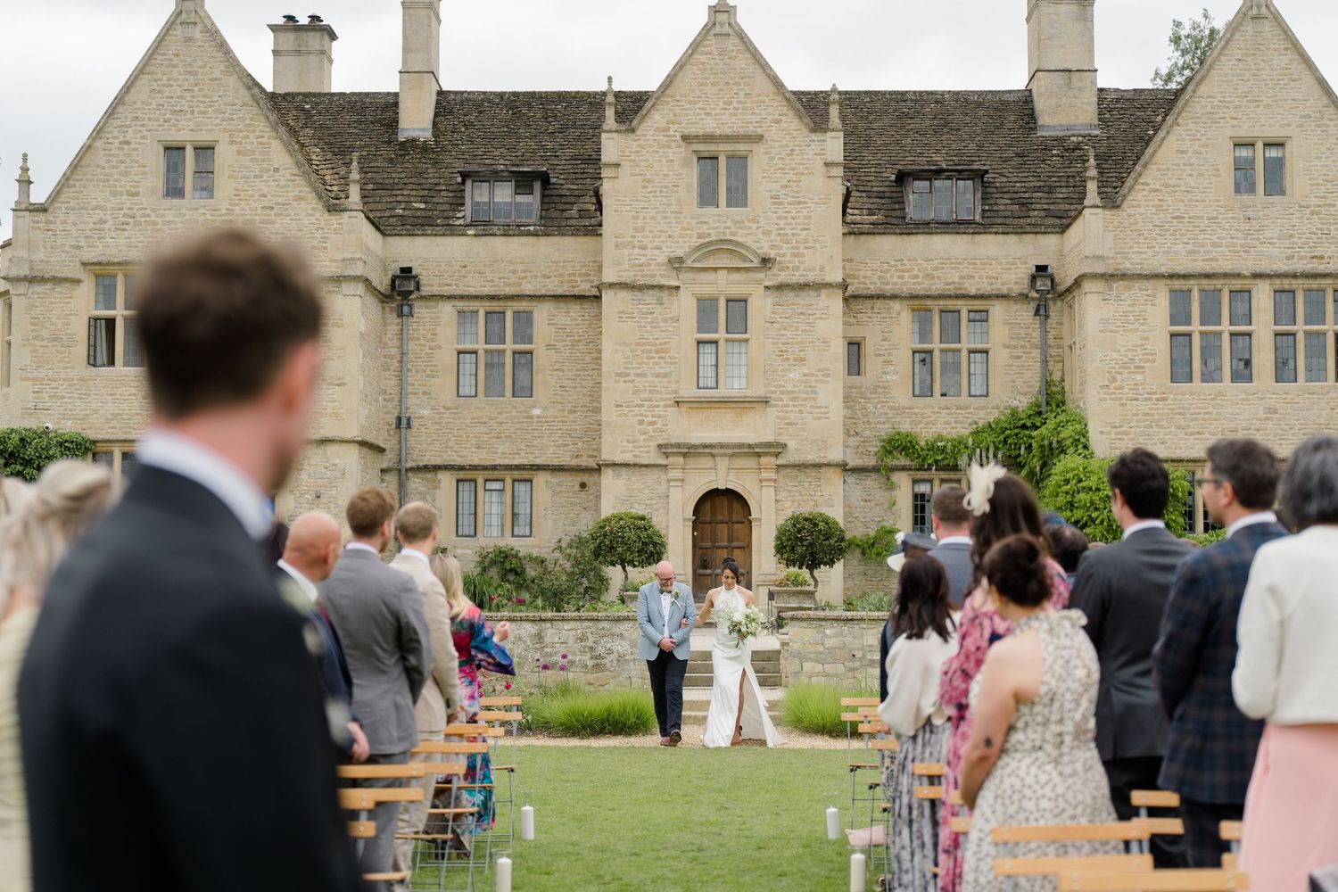 Outdoor wedding ceremony on the lawn of a grand English stone manor house with seated guests.