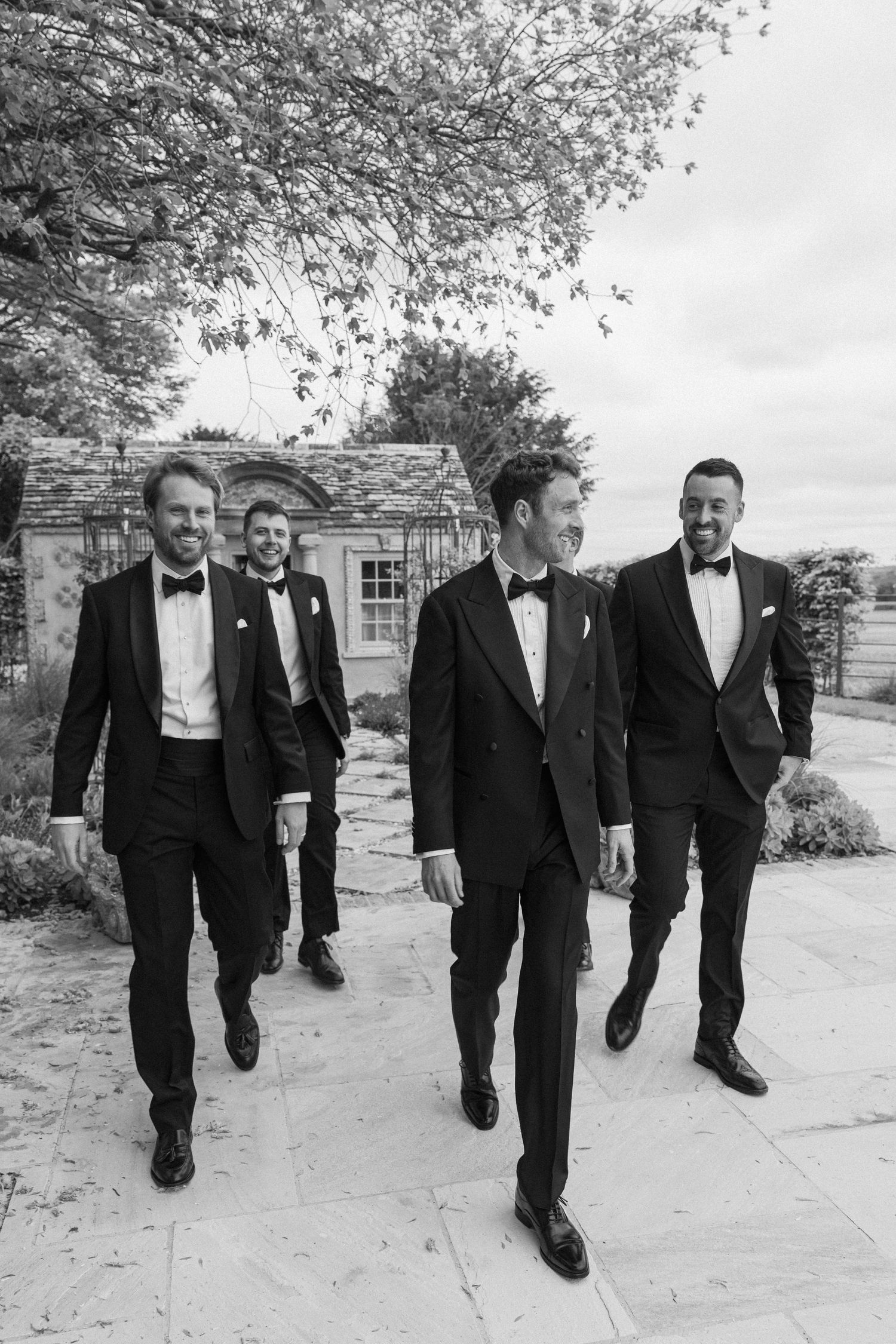 Four groomsmen in black tuxedos walking confidently together outside a stone wedding venue.