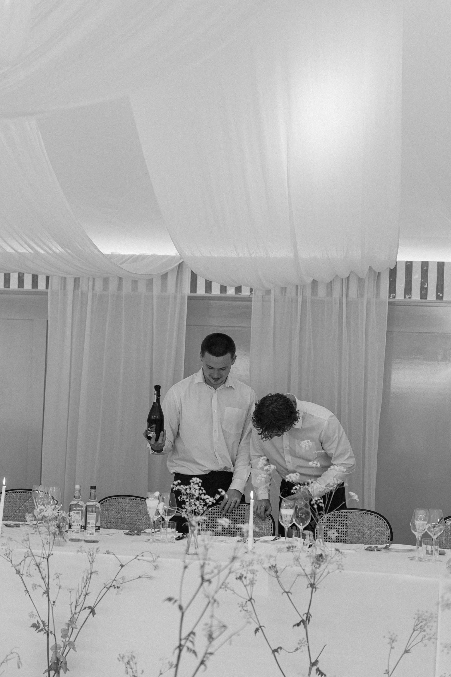 Two waitstaff prepare drinks at a wedding reception table beneath draped white marquee ceiling in black and white.