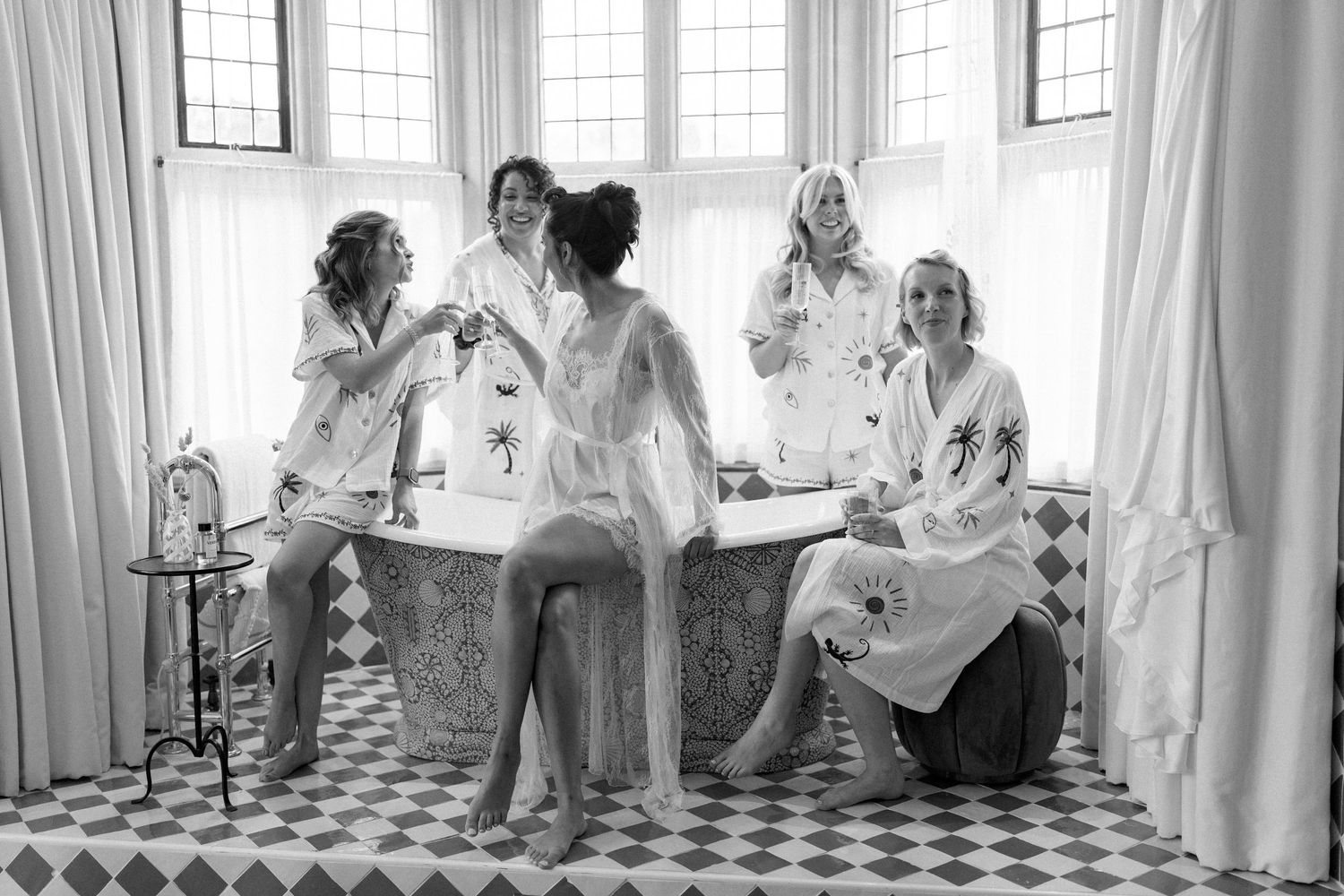 Bridesmaids in matching robes celebrating around clawfoot bathtub in black and white photo.