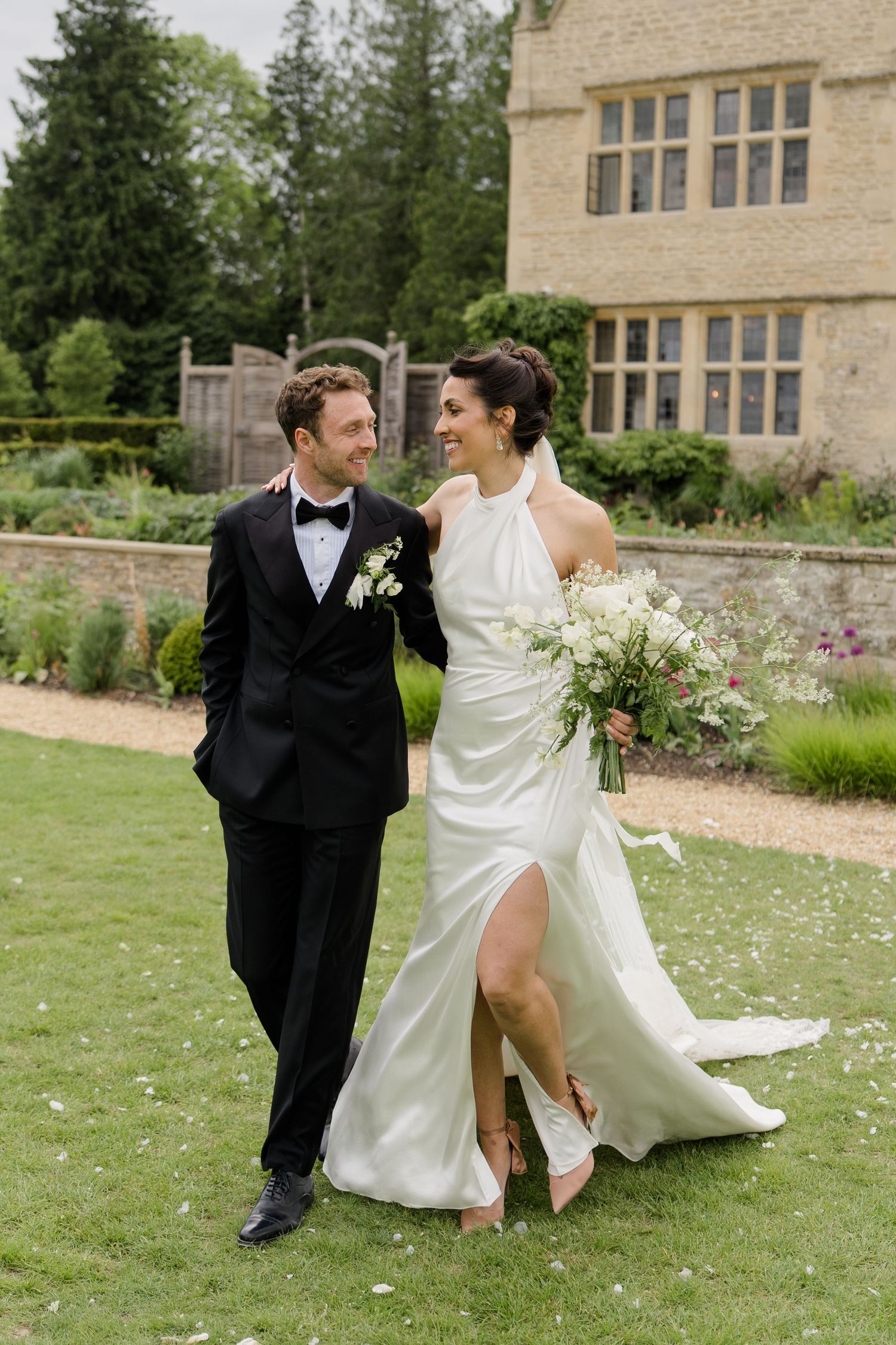 Smiling bride in halter neck gown with slit and groom in tuxedo walk across lush green lawn.