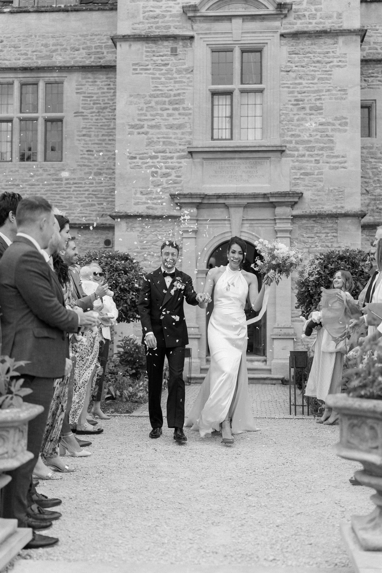 Bride and groom walking through confetti outside stone castle wedding venue in black and white.