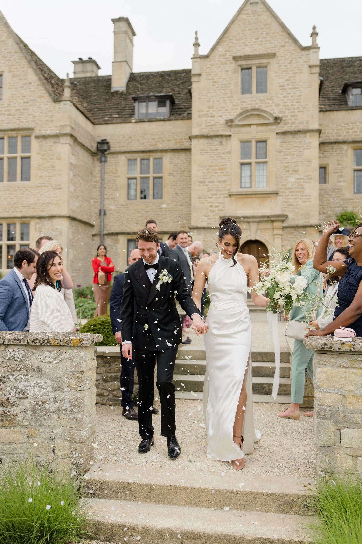 Bride and groom walk through confetti shower outside a stunning Cotswolds stone manor house wedding venue.