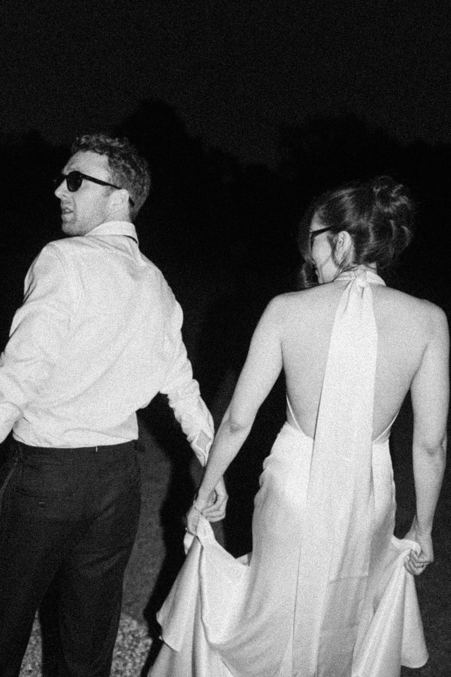 Bride and groom in sunglasses walk hand in hand into the night, elegant backless wedding gown detail.