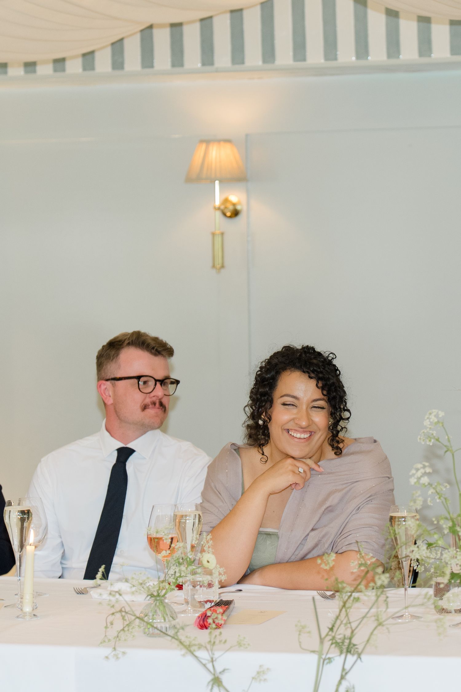 A smiling woman with curly hair and a man in glasses enjoy a wedding reception dinner together.