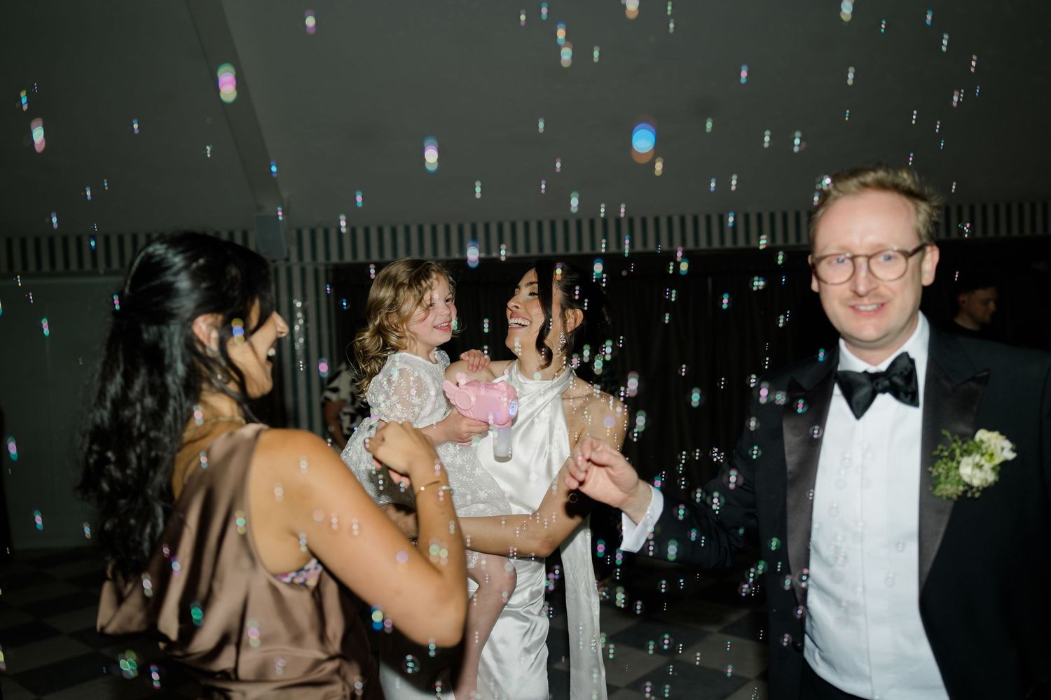 Bride dancing joyfully with guests at wedding reception with bubbles floating in the air.