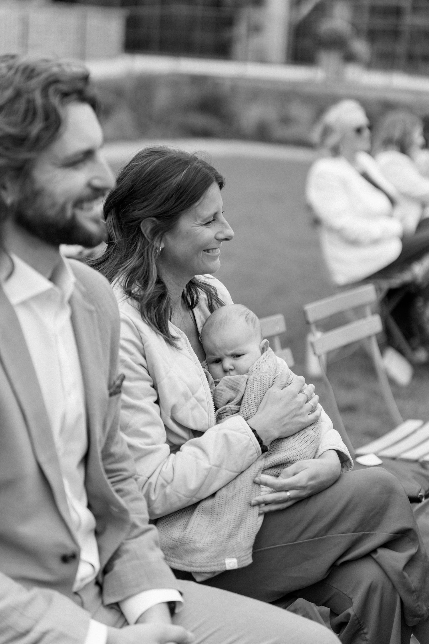 Black and white photo of smiling wedding guests seated outdoors, with a woman cradling a baby.