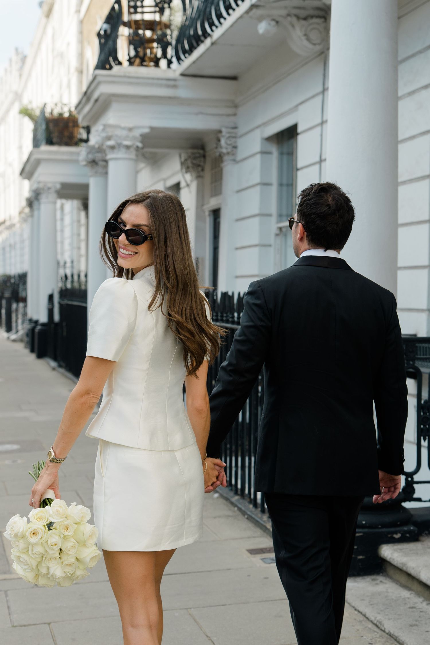 Smiling bride in white dress with bouquet walks hand-in-hand with groom along white townhouse street.
