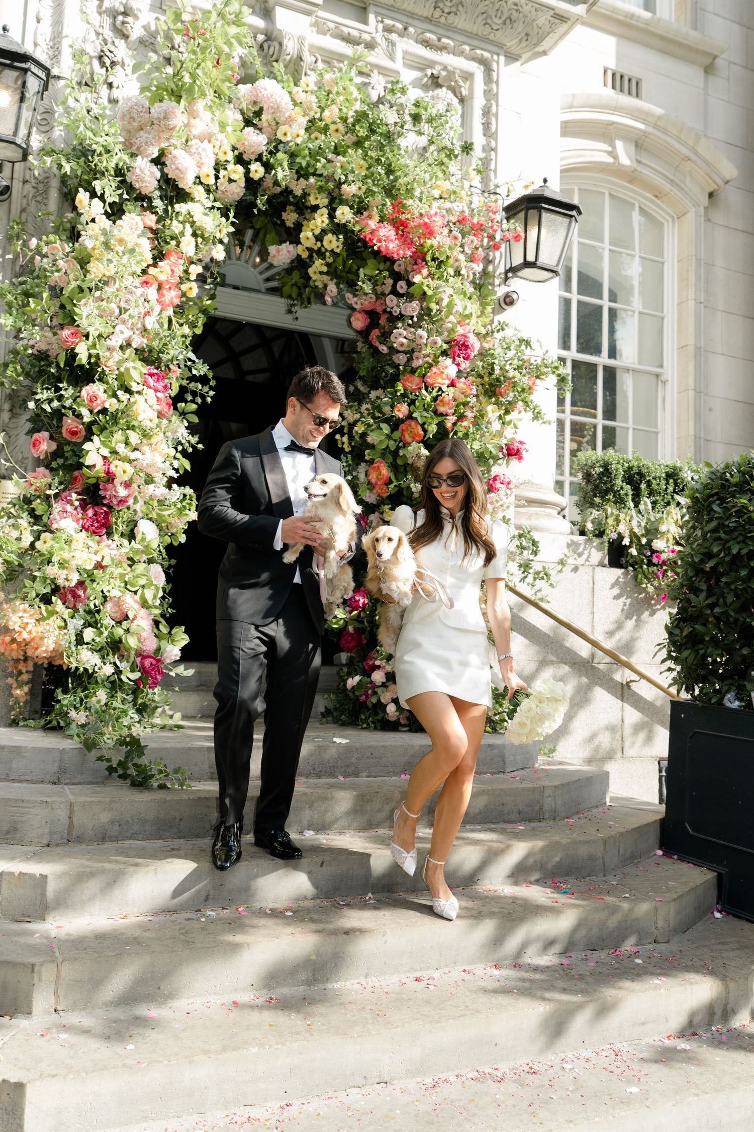 Newlyweds carrying dogs descending steps beneath stunning floral arch of pink and white blooms.