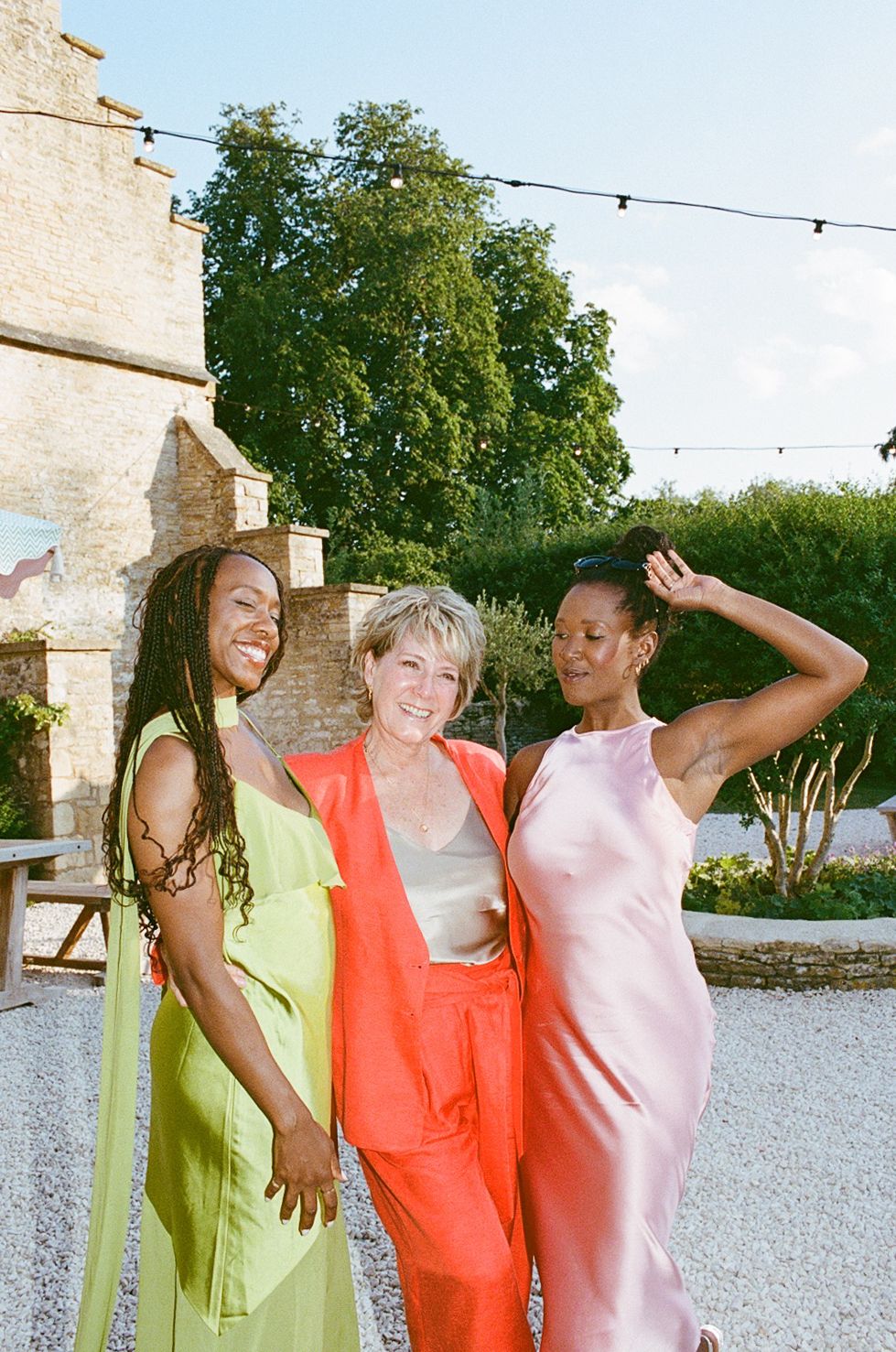Three women in vibrant outfits smile outdoors at a garden event, wearing lime green, orange, and pink dresses.