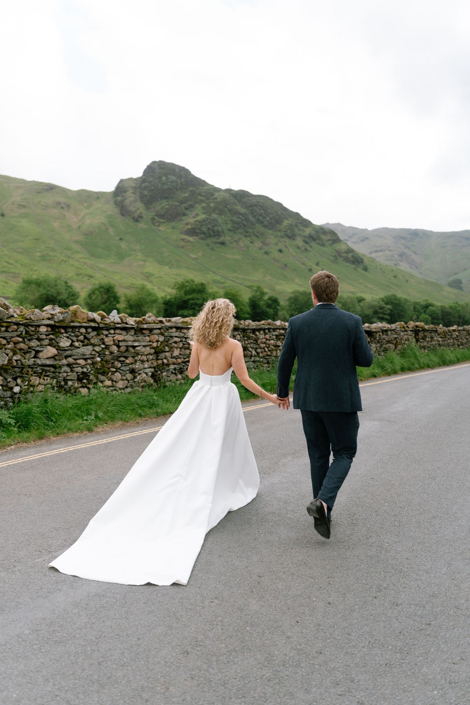A couple walks away from camera on a country road with mountains and stone walls in background.