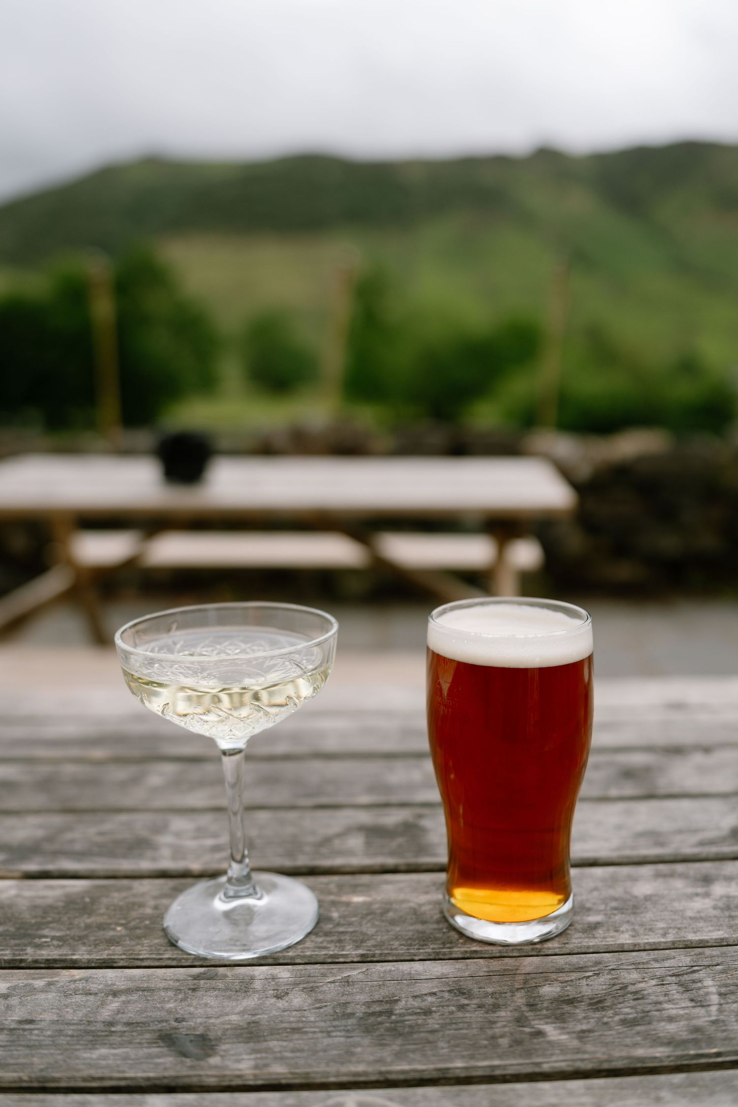Two drinks sit on a weathered wooden table with rolling hills in the background.