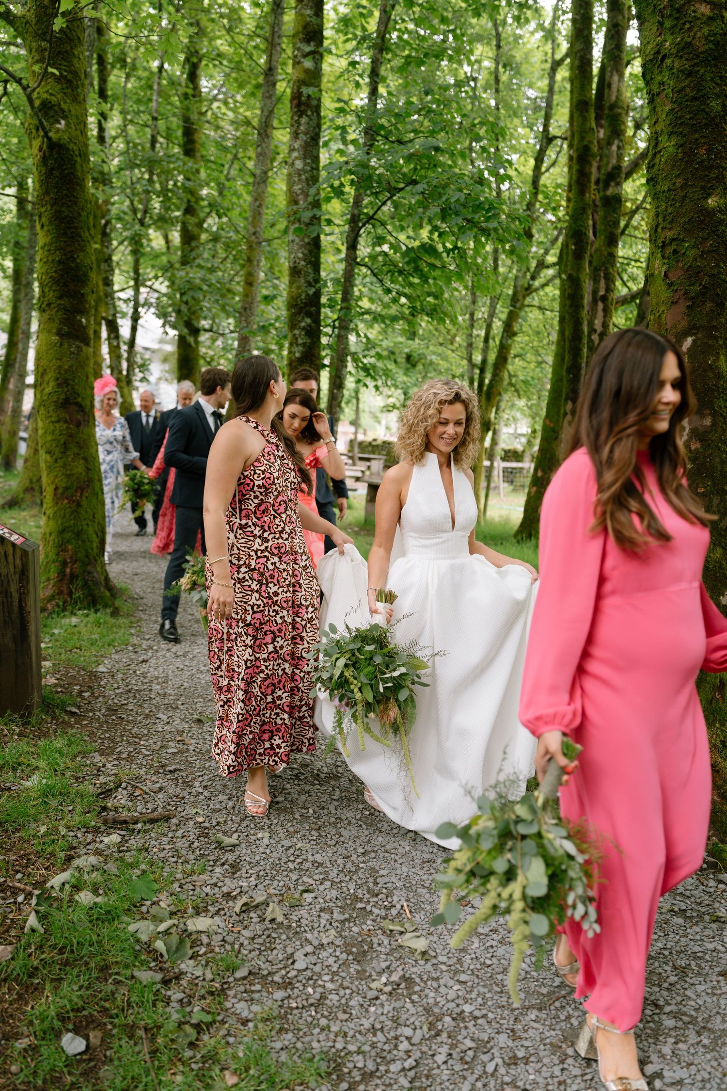 Wedding party walks along a wooded path with bridesmaids in coral dresses and a bride in white.