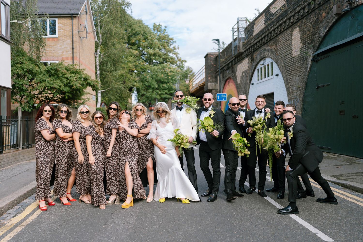 Wedding party poses creatively for group photos on an urban street.