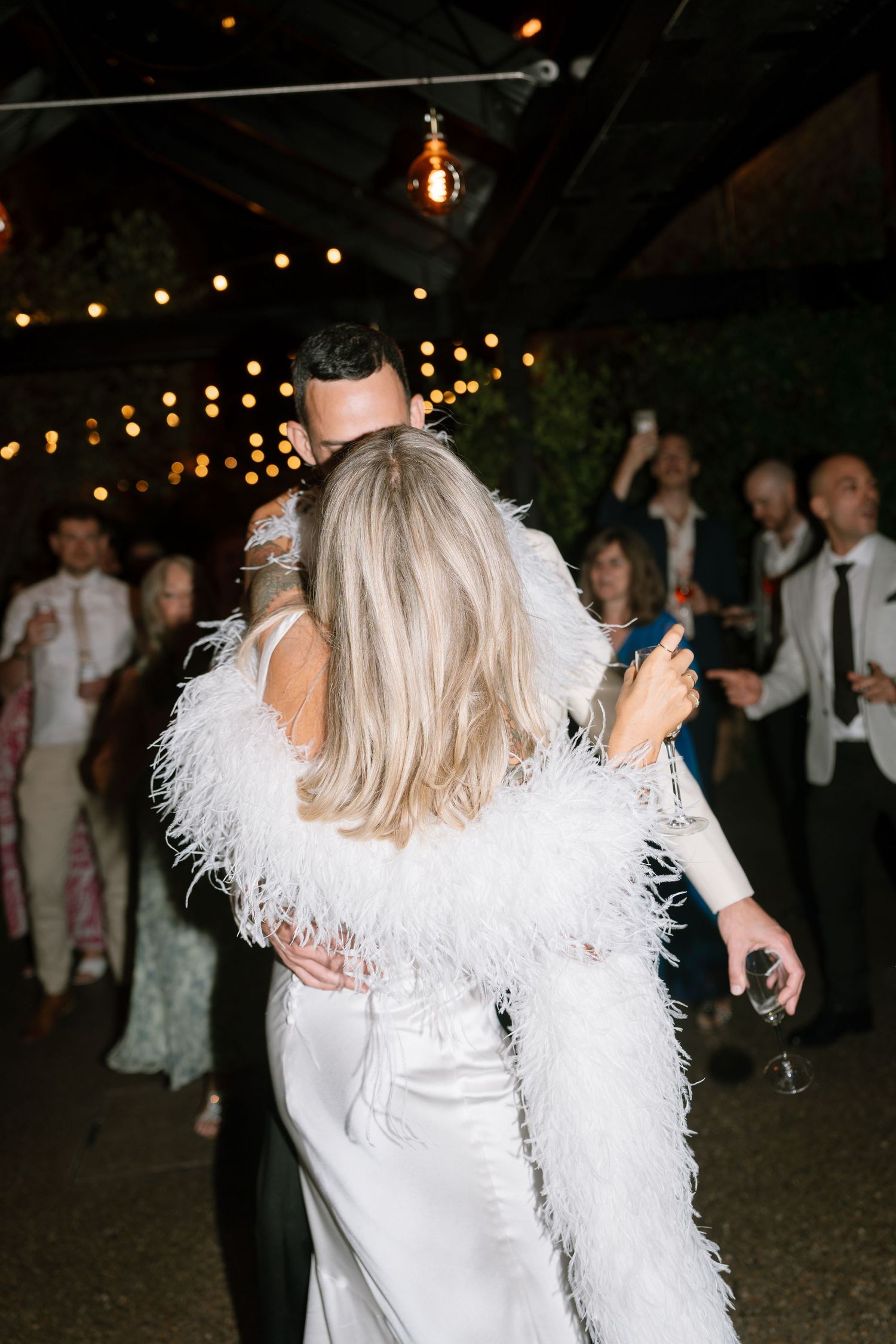 Couples dance under twinkling lights at a wedding reception, with one wearing a white feathered shawl.